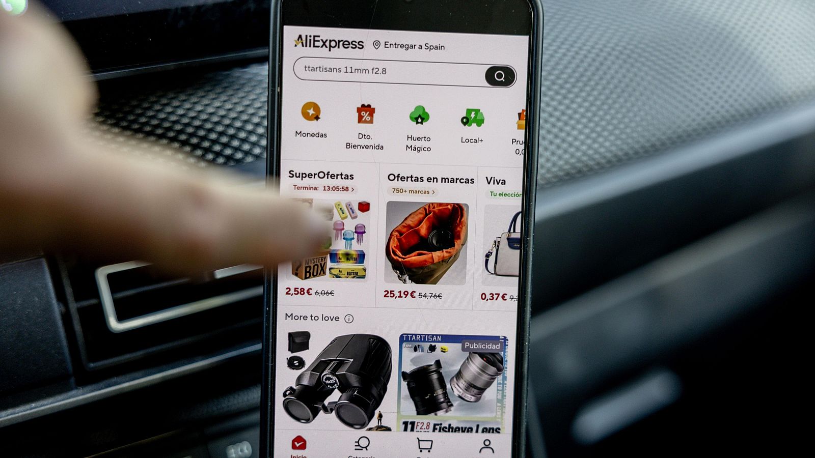 Primer plano de un móvil con la app AliExpress, mostrando ofertas y productos. Una mano interactúa con la pantalla. Fondo de vehículo.