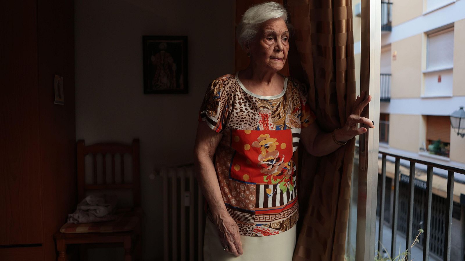 Paquita, de 82 años, mira por la ventana con preocupación. Lleva una blusa estampada y falda clara. El interior está oscuro, el exterior muestra edificios y una farola.