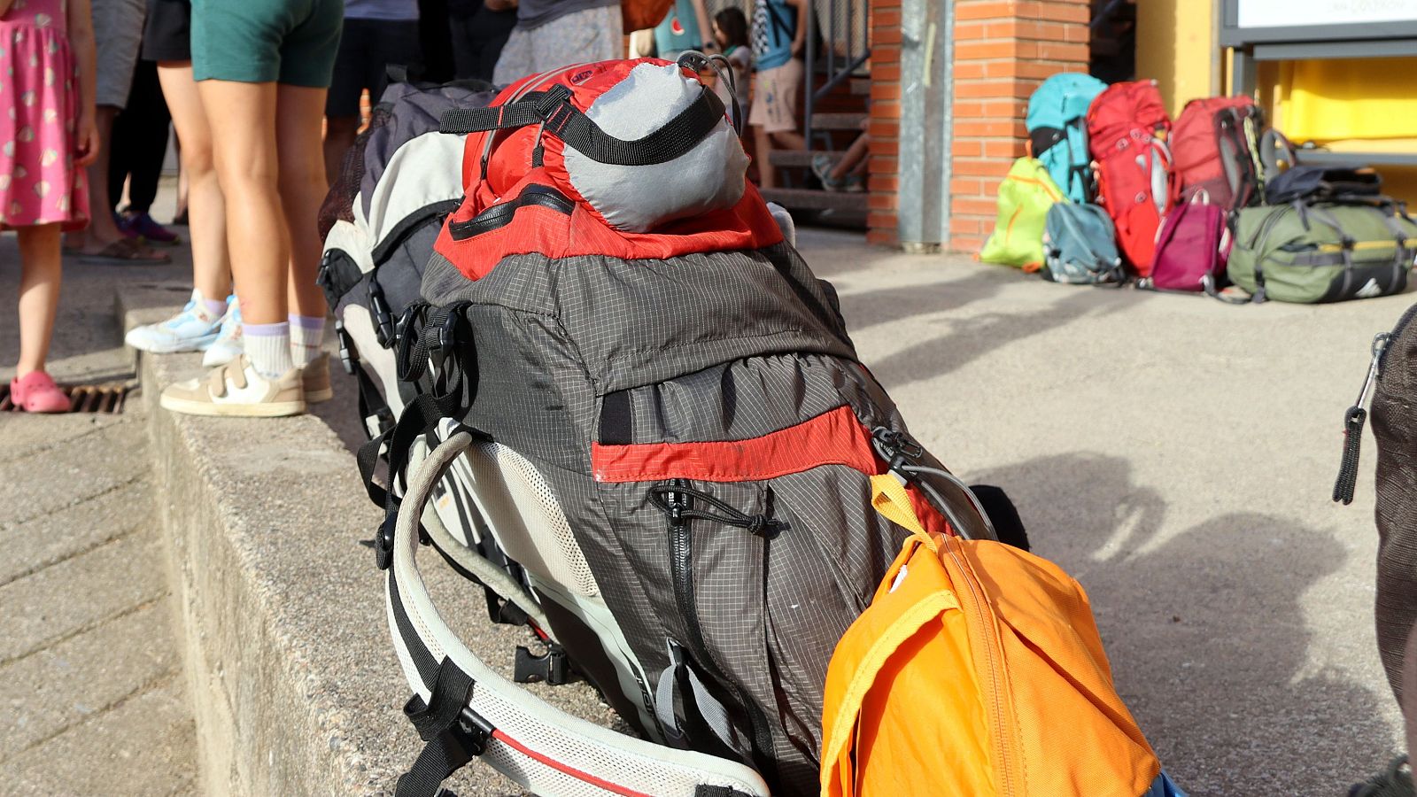 Mochilas de senderismo de varios colores y tamaños, junto a las piernas de personas, algunas con ropa de verano, en un día soleado.