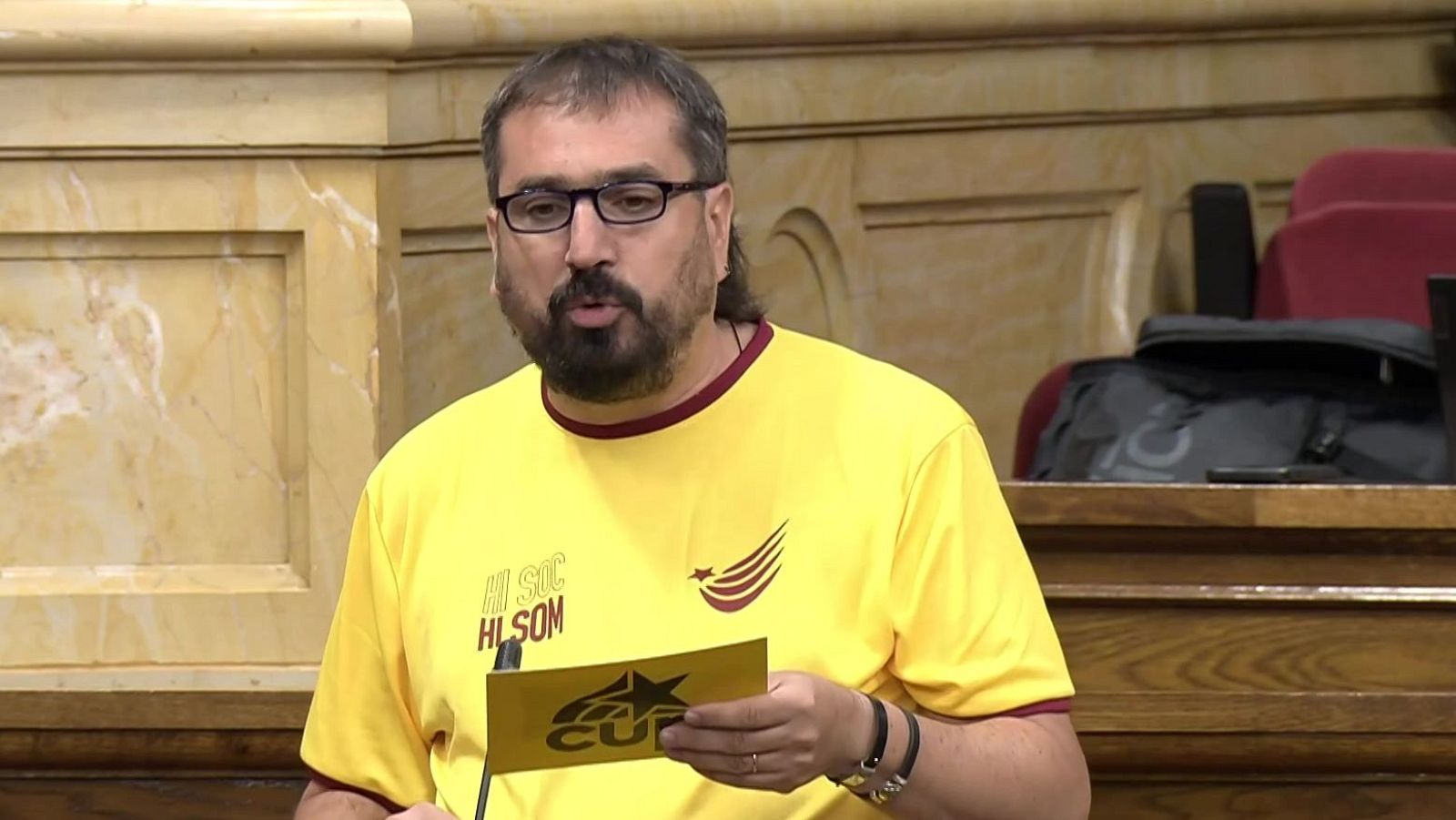 Primer plano de Dani Cornellà, diputado de la CUP, con camiseta amarilla "HI SOM" y logo, sosteniendo tarjeta CUP. Expresión de habla.