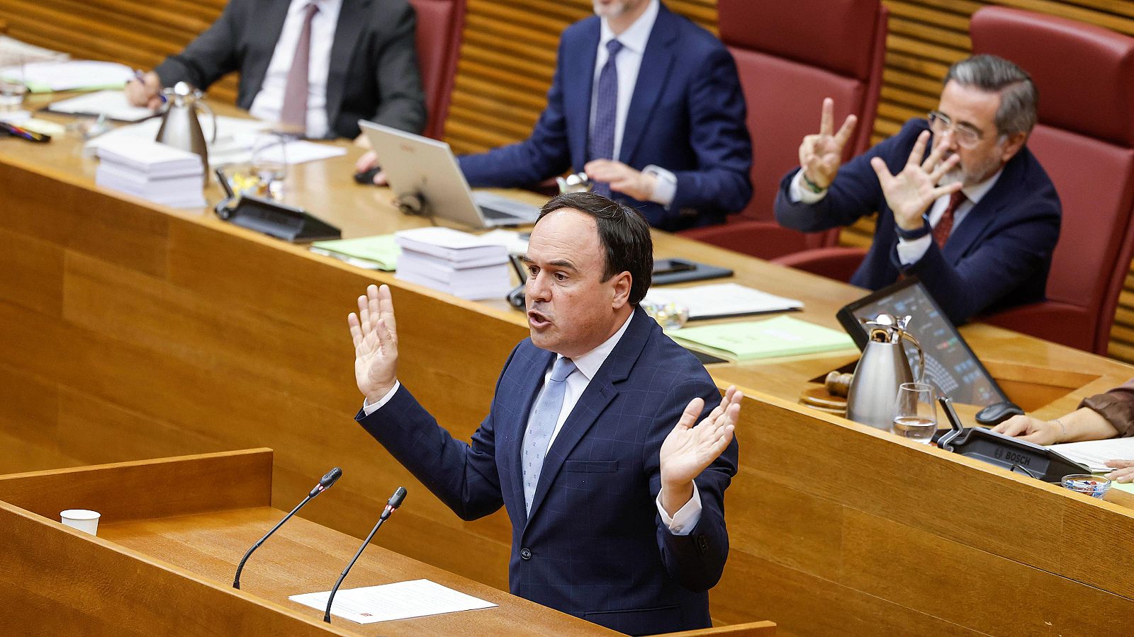 Juanfran Pérez Llorca, de pie en un atril, pronuncia un discurso con gestos enfáticos. A su derecha, un hombre con gafas y barba gesticula activamente. Ambos participan en una sesión plenaria.