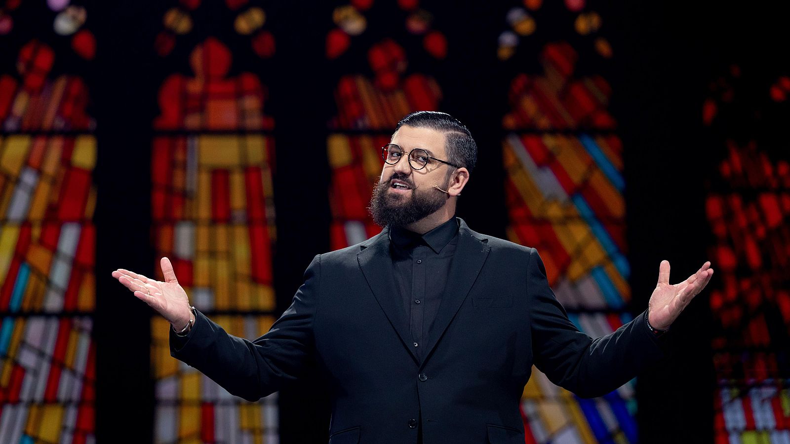 Manu Sánchez, con traje oscuro y gafas, gesticula con los brazos extendidos ante un fondo de vidrieras de colores.