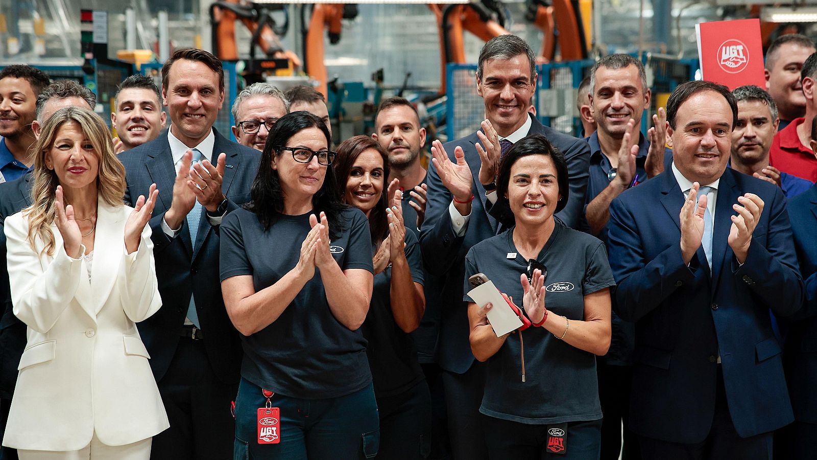 Pedro Sánchez, Yolanda Díaz y Jim Baumbick celebran en una planta de Ford rodeados de maquinaria industrial y otros asistentes.