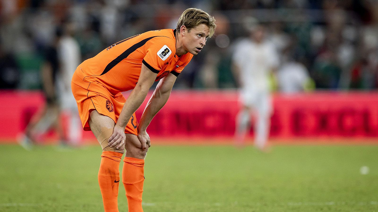 Frenkie de Jong, con la camiseta naranja de Países Bajos (número 21), se inclina en un campo de fútbol, concentrado. Fondo con valla publicitaria y siluetas.