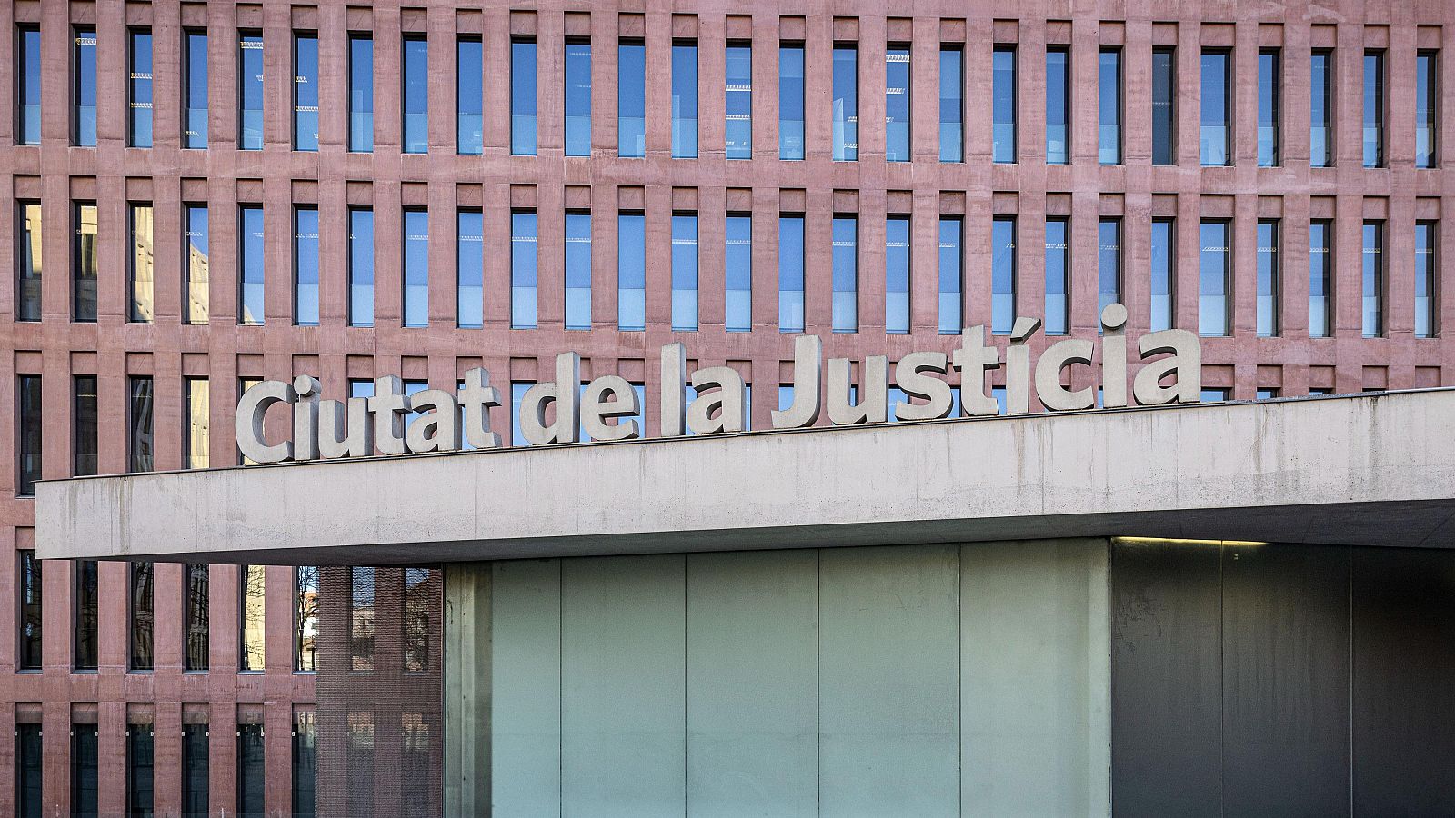 Vista frontal de la Ciutat de la Justícia de Barcelona, amb façana de finestres marrons vermelloses, estructura de formigó gris i el nom del edifici en lletres blanques.