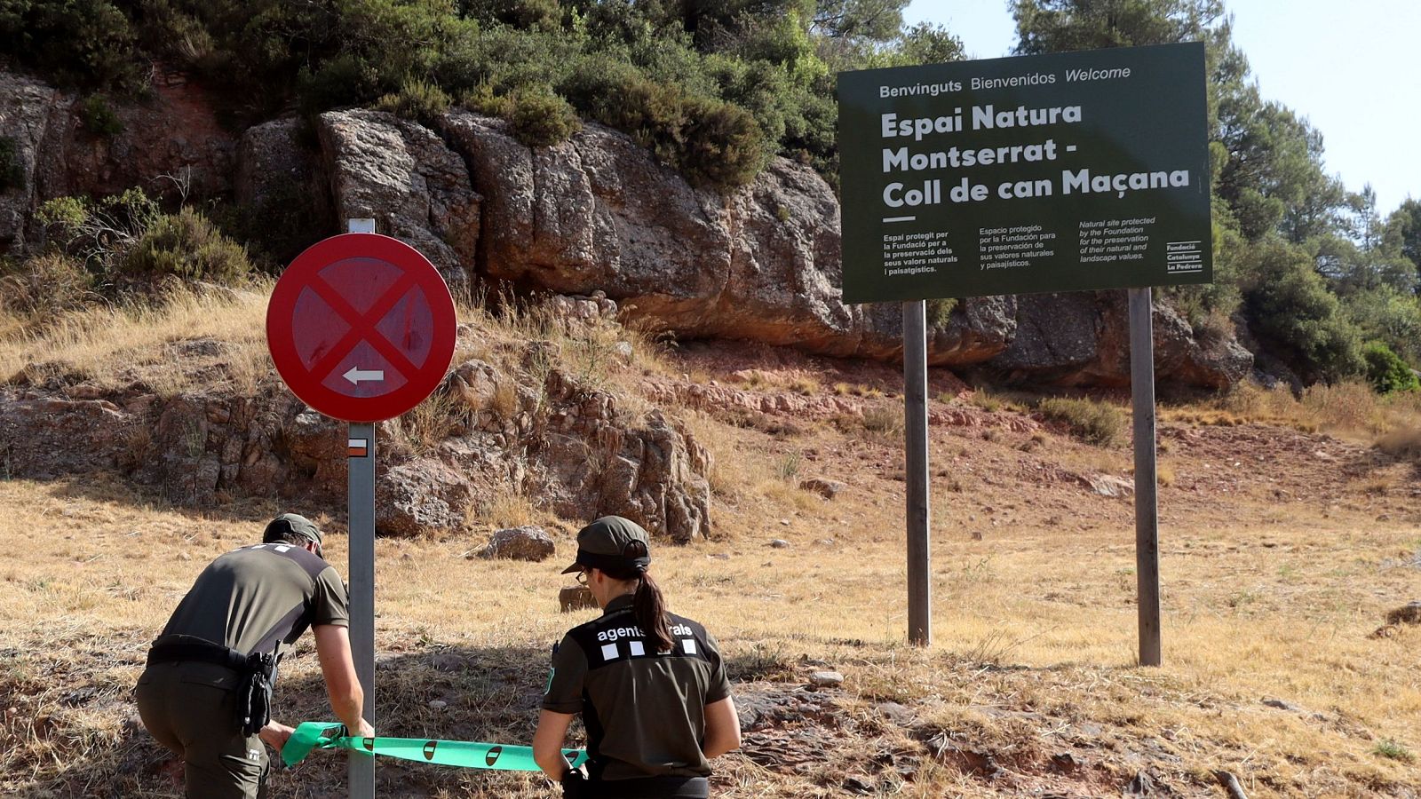 Agents rurals amb uniformes verds restringeixen l'accés al Coll de can Maçana, Espai Natura Montserrat, amb una cinta i una senyal de prohibició. Un cartell dona la benvinguda.
