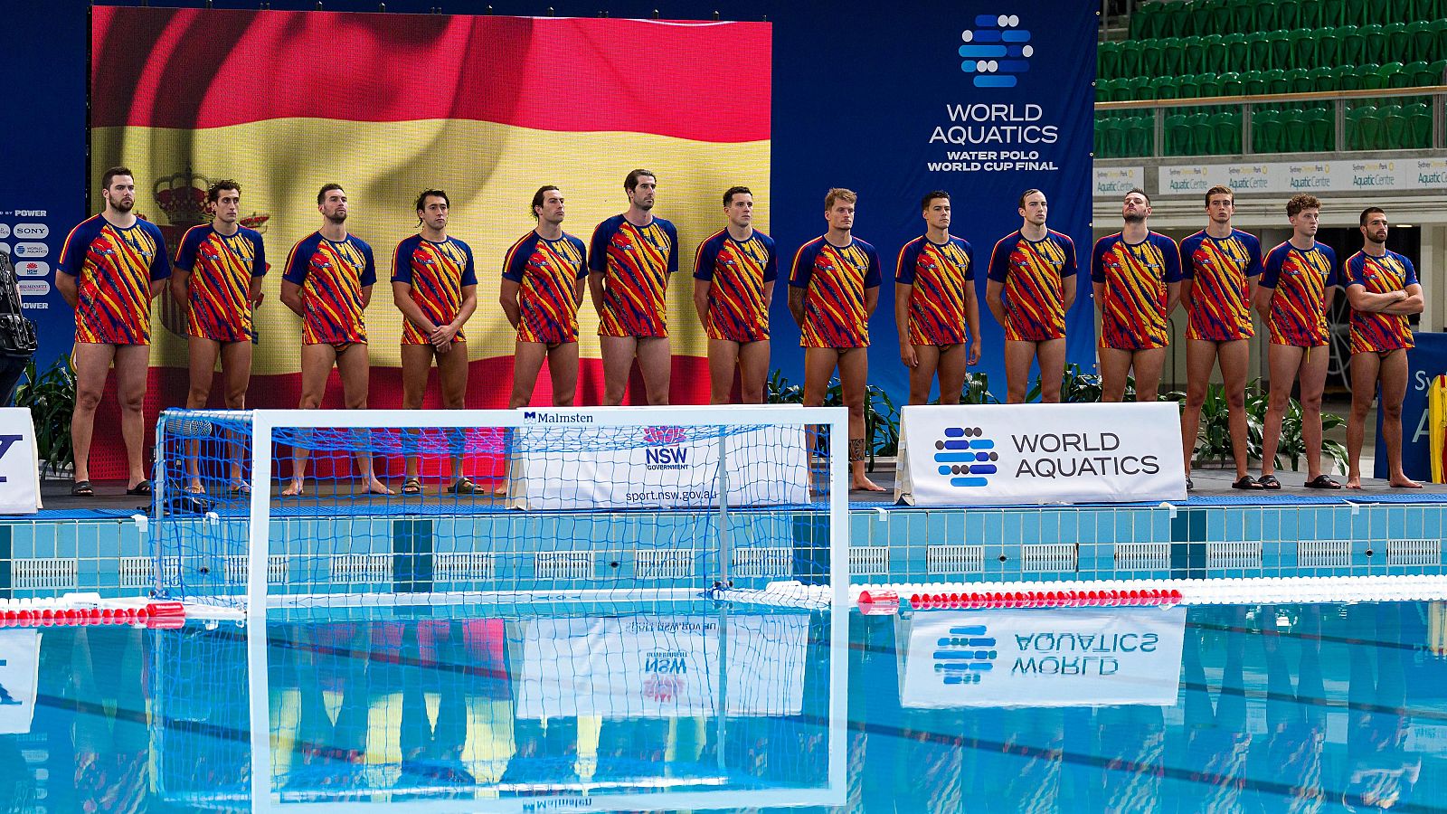 Jugadores de waterpolo de España, con camisetas de rayas rojas, amarillas y naranjas, posan ante una bandera de España y el logo de "WORLD AQUATICS" en la "WATER POLO WORLD CUP FINAL".