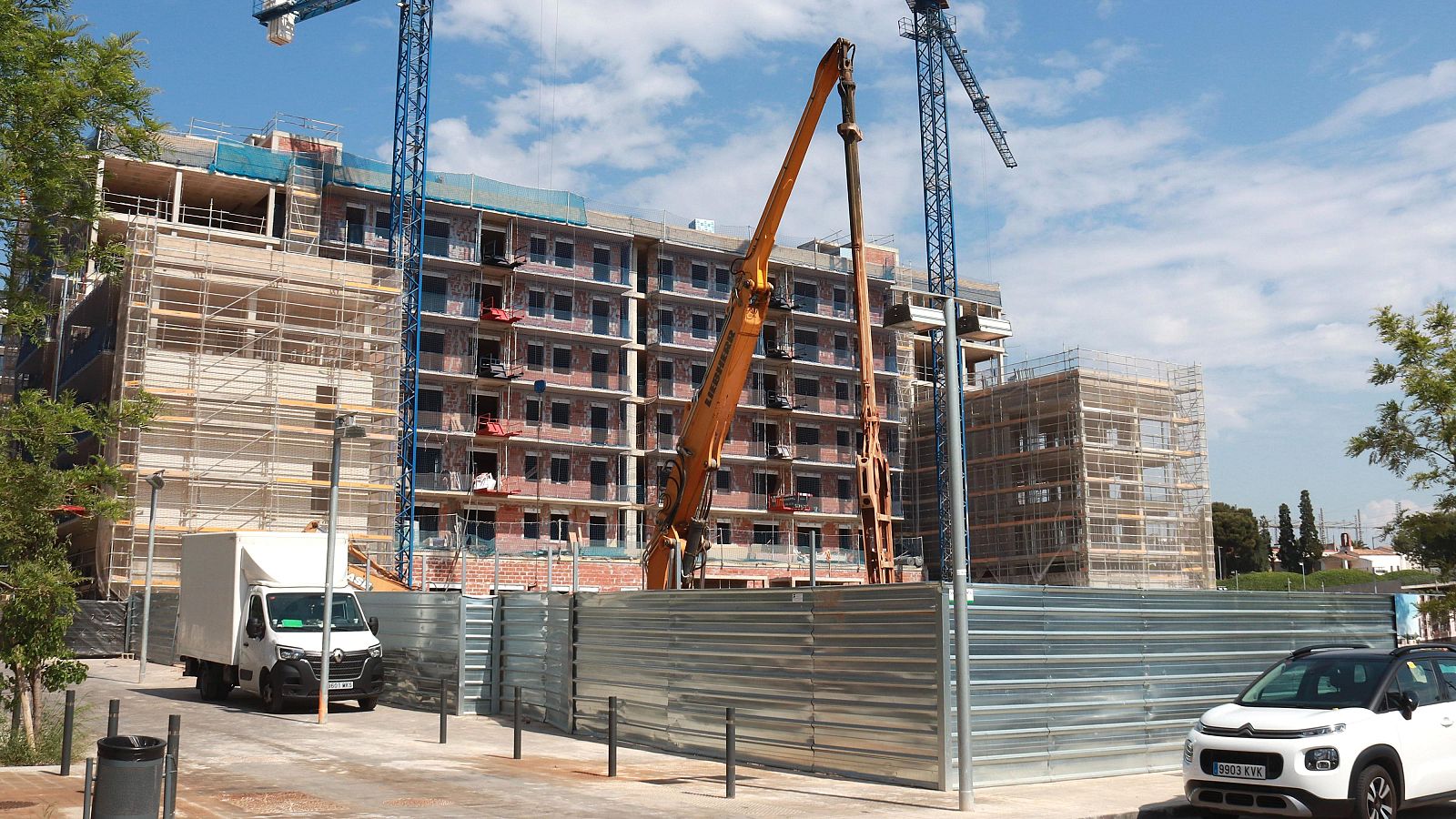 Amplio solar en construcción de viviendas con grúas, maquinaria Liebherr y edificios en distintas fases. Furgoneta Renault y Citroën C3 Aircross estacionados en primer plano.