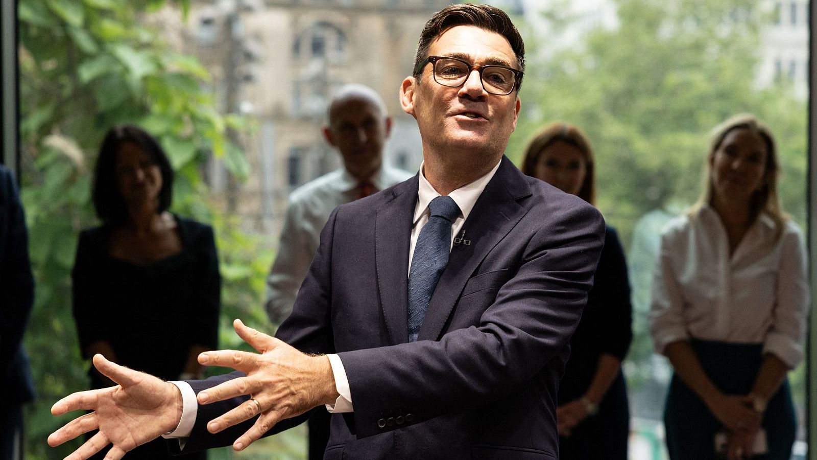 Andy Burnham, Alcalde de Gran Manchester, habla en un evento formal. Viste traje azul marino y gafas, con las manos extendidas en un gesto expresivo. Al fondo, se ve a la audiencia y ventanales.