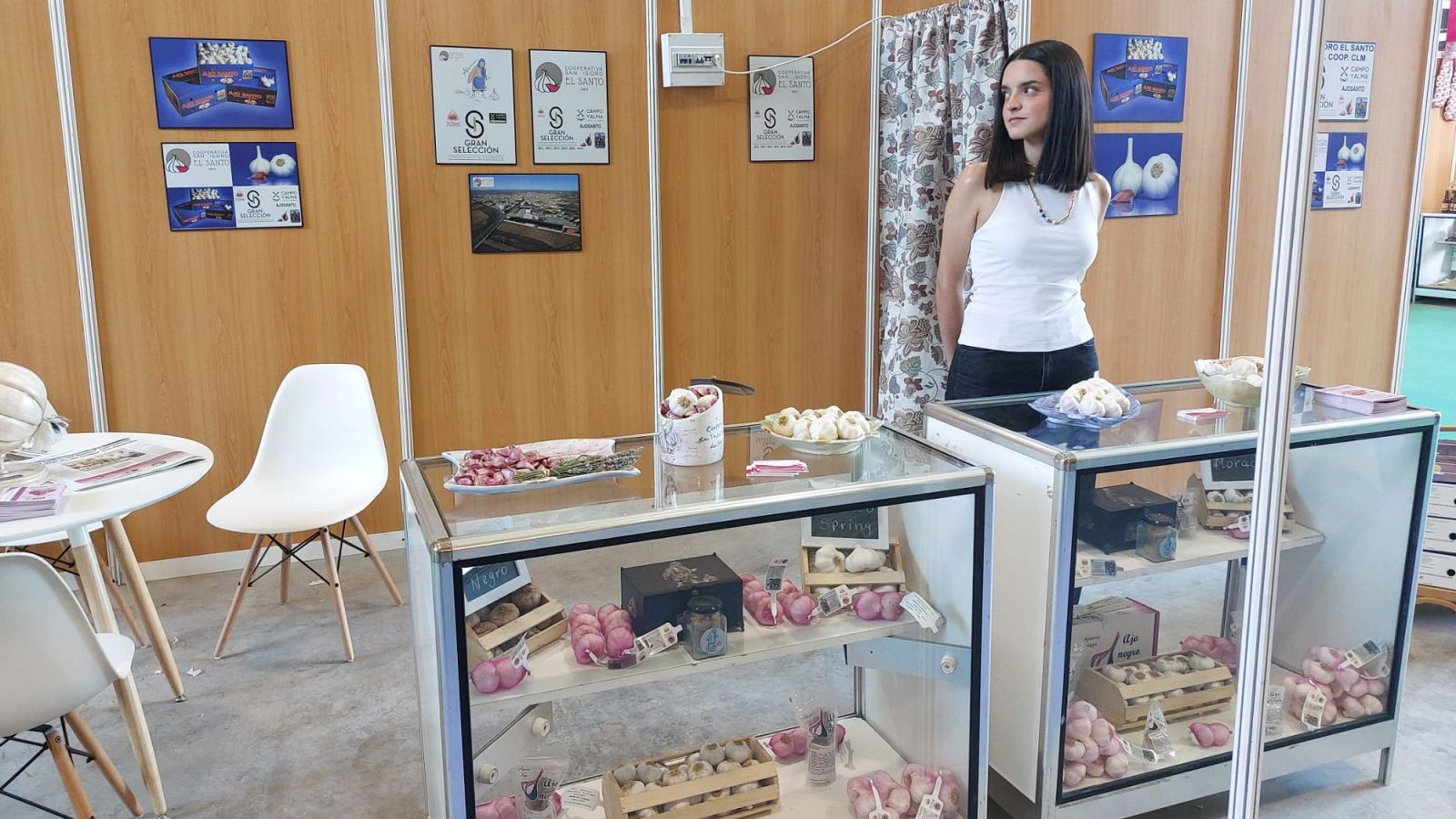 Stand de exposición con vitrinas de ajo, pósteres y una mujer joven. Se observan ajos morados, negros y productos derivados en envases variados. Probable evento en Las Pedroñeras.