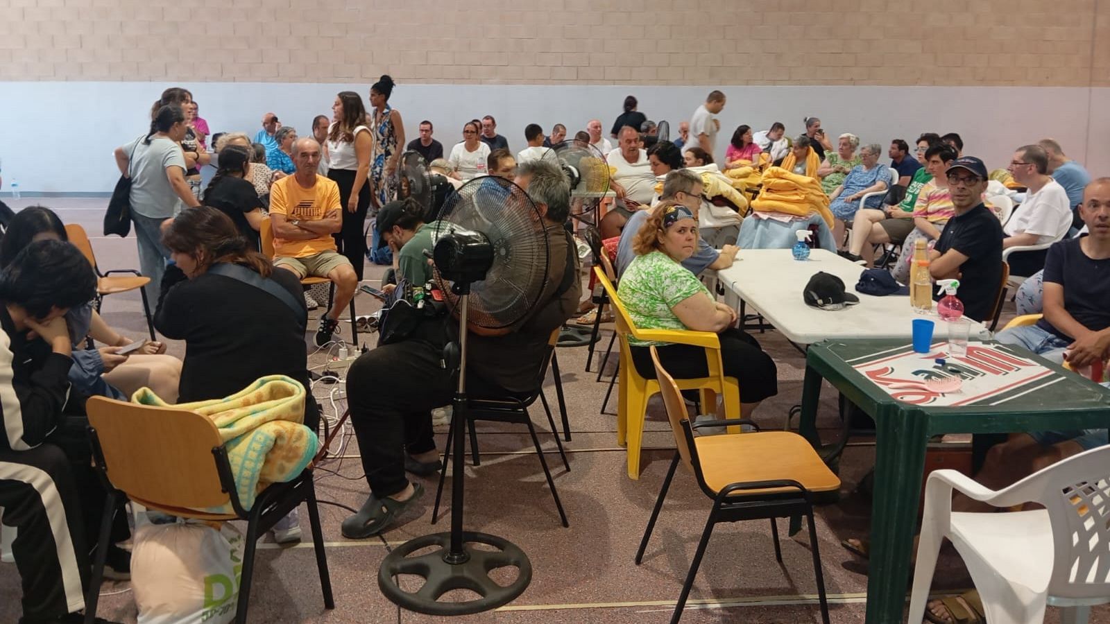 Decenas de personas, mayormente adultos y mayores, se refugian en un pabellón interior, sentados en sillas y mesas, con ventiladores y pertenencias a la vista.