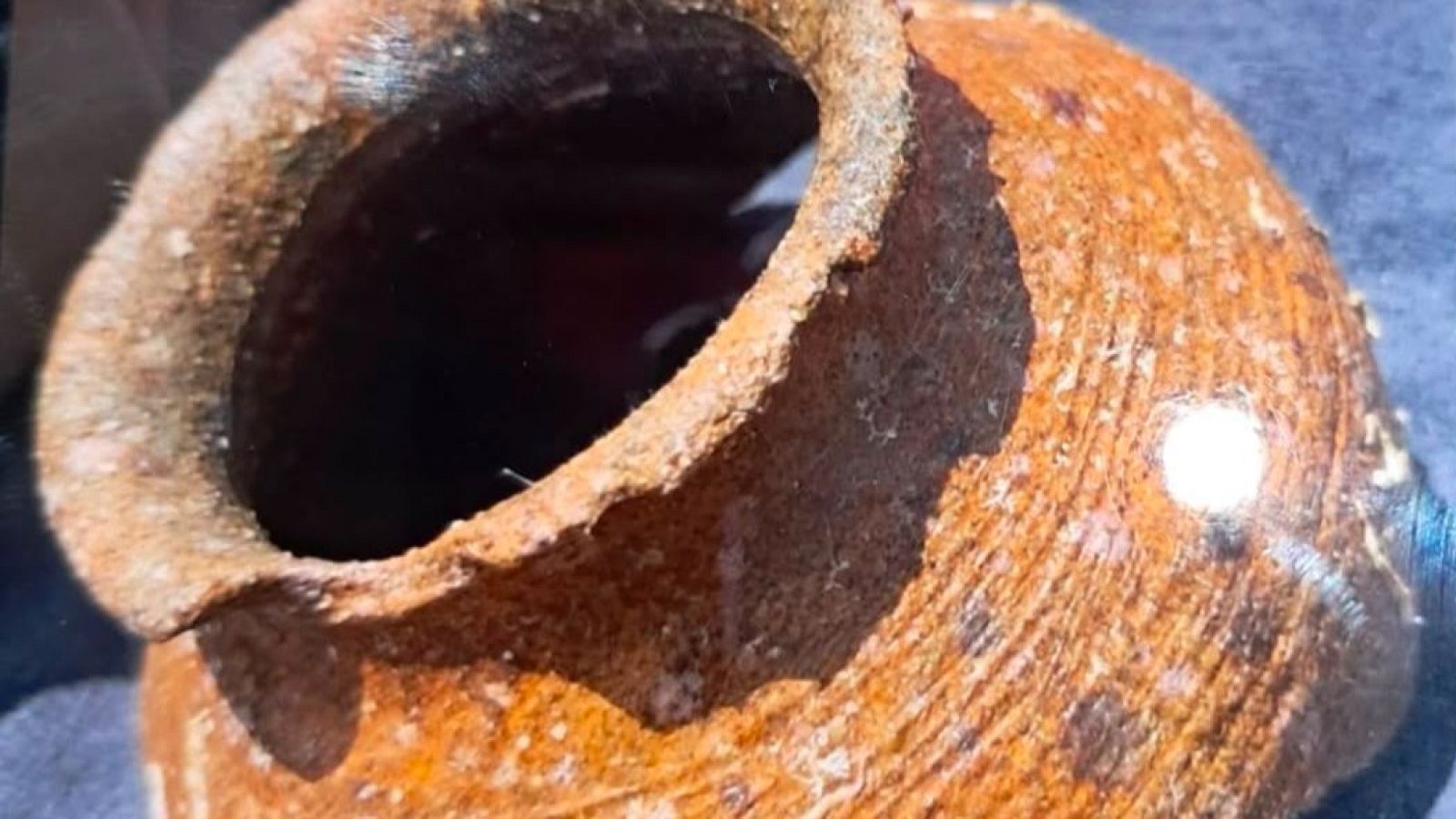 Vasija antigua de tonos terrosos, textura rugosa y borde irregular, con un reflejo de luz en su superficie.