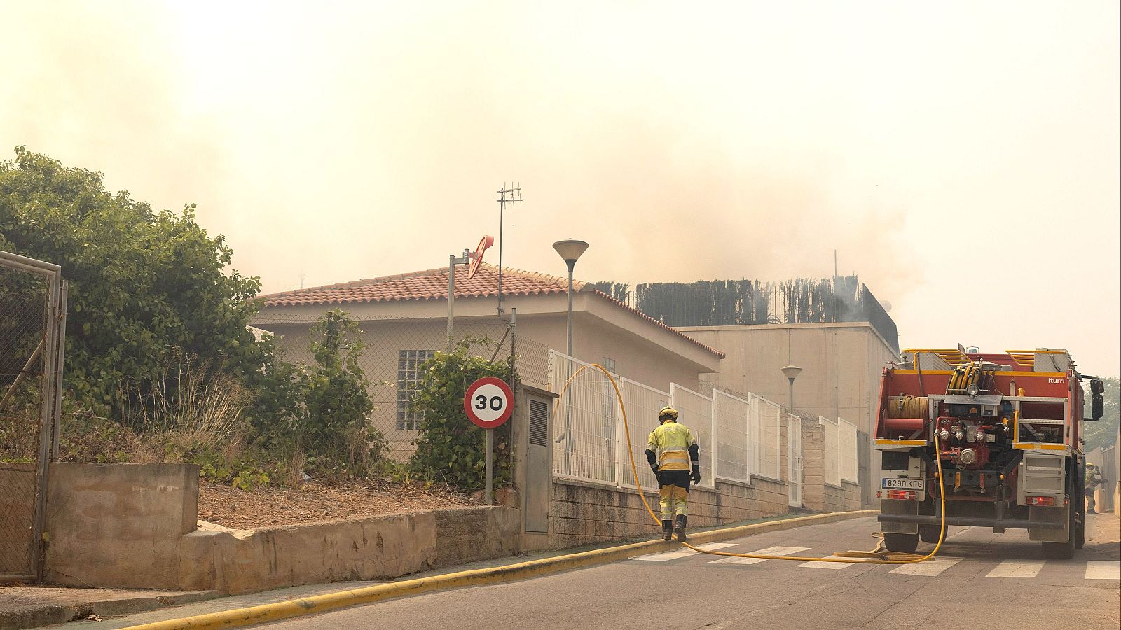 Bombero arrastra manguera amarilla de camión de bomberos rojo y amarillo bajo cielo teñido de humo por incendio.