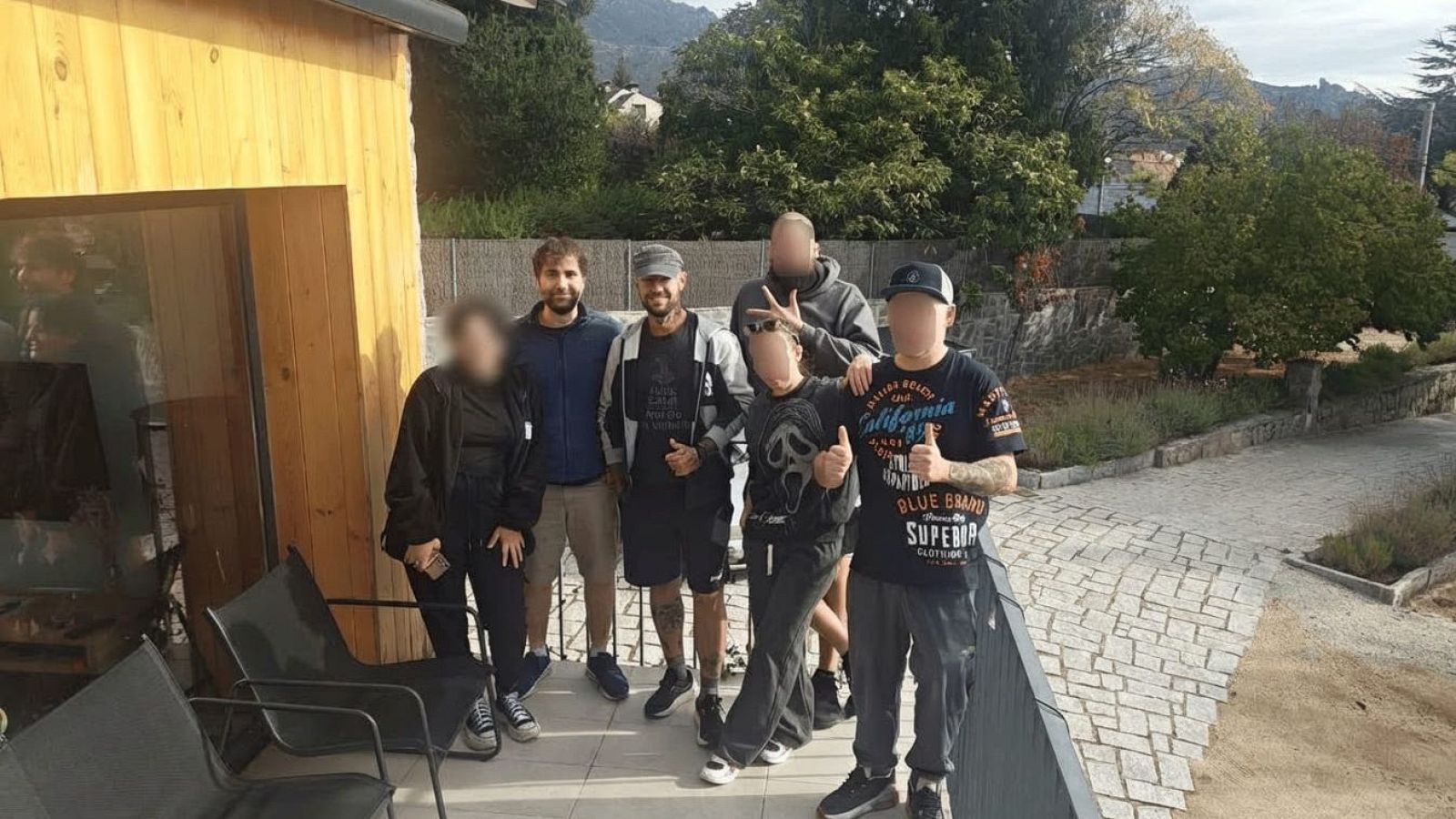 Seis personas, incluyendo a Pablo Iglesias y Pablo Hasél, posan en una terraza con montañas al fondo. Se aprecian detalles de vestimenta, tatuajes y gestos como pulgares hacia arriba y cuernos.