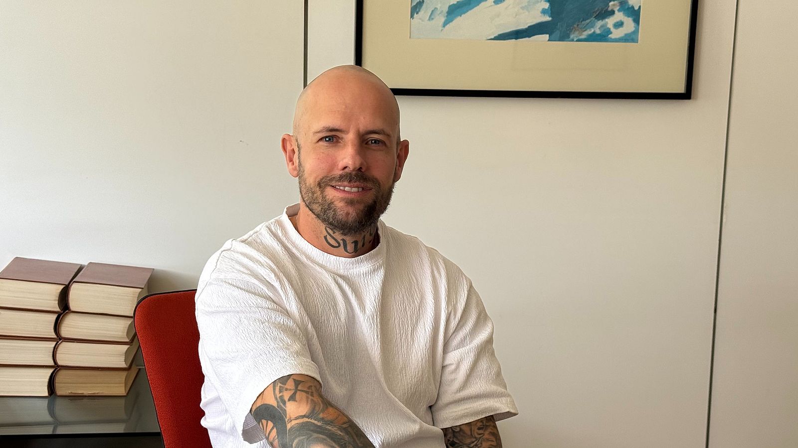 Retrato de David Saavedra, hombre calvo con barba y bigote, sonriendo a la cámara con tatuajes visibles, camiseta blanca, fondo blanco con cuadro y libros.