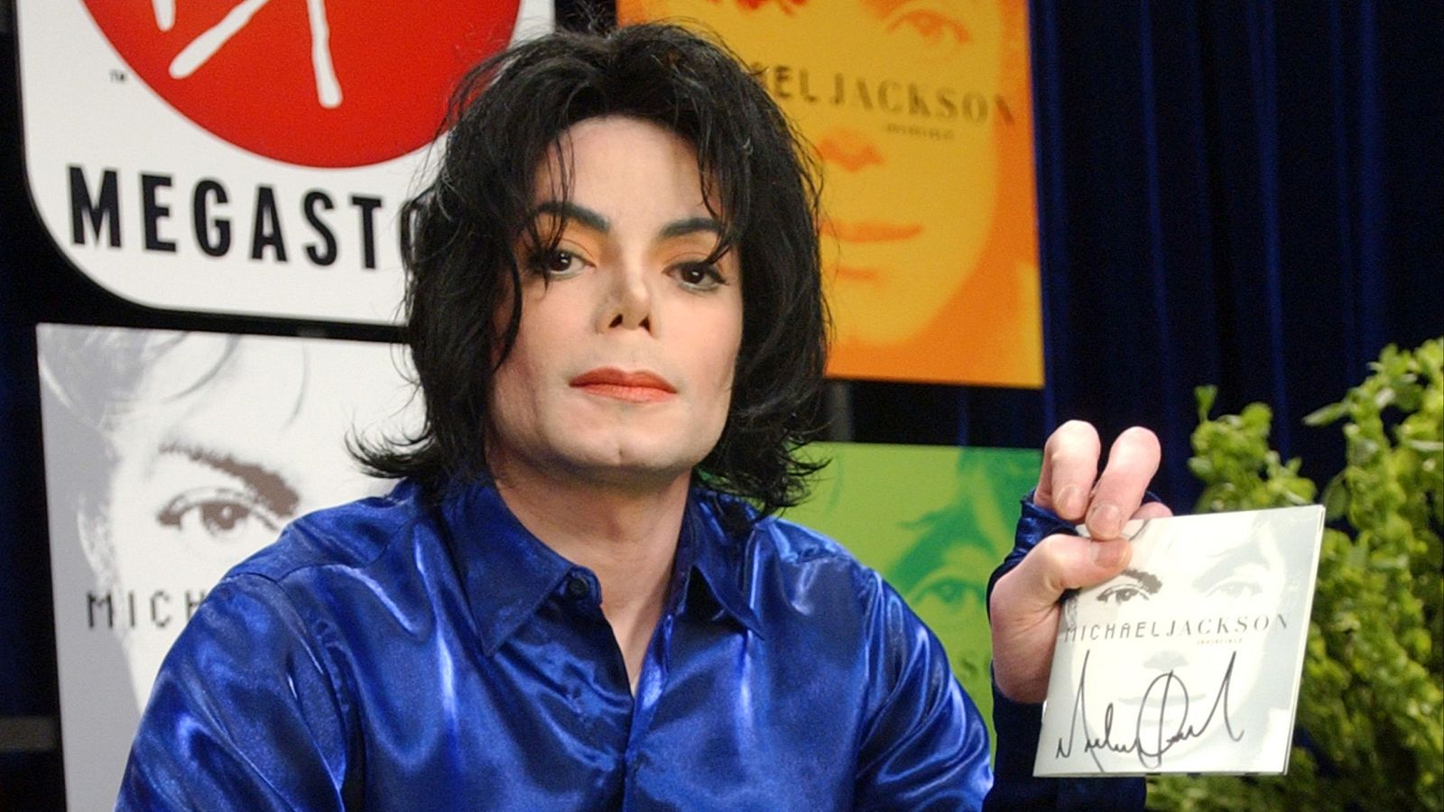 Michael Jackson promociona su álbum "Invincible", sosteniendo la carátula con su nombre y firma, rodeado de material promocional del disco.