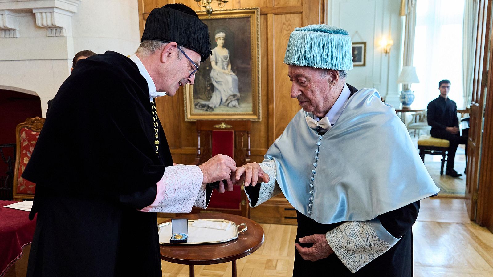 Ceremonia académica: el rector Ángel Pelayo impone un anillo a José María Pérez Peridis como doctor "honoris causa", ambos con togas académicas, en un salón con paneles de madera y un retrato.