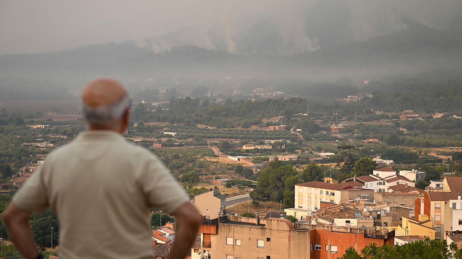 Hombre mayor observa Onda (Castellón) envuelta en humo de incendio forestal. Paisaje urbano y montañoso difuminado por la bruma grisácea.