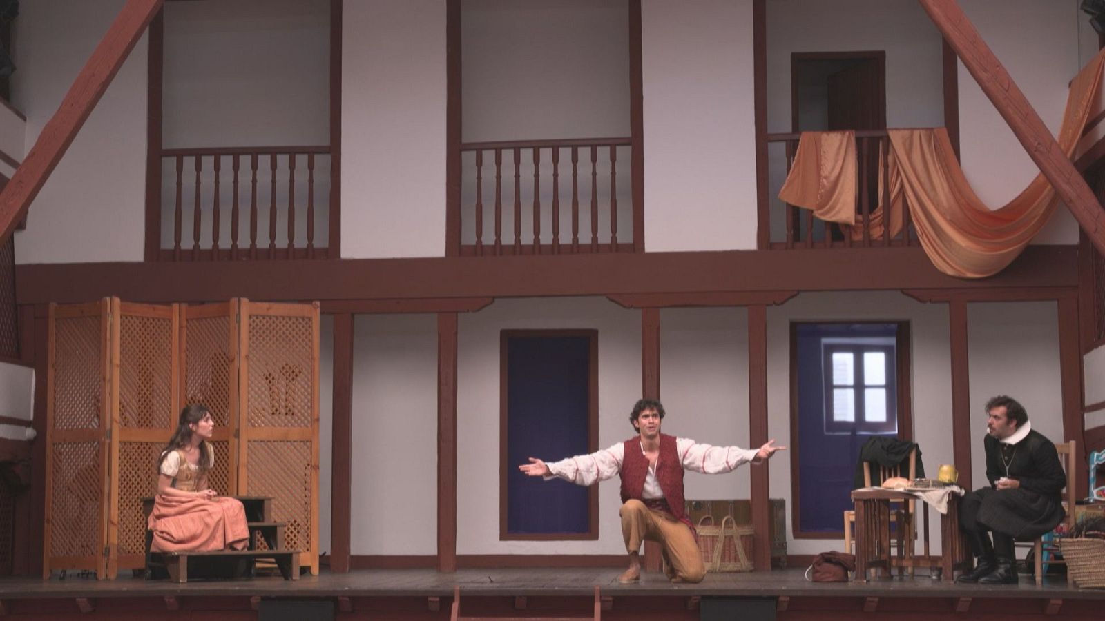 Escena teatral con arquitectura clásica española. Un hombre arrodillado, una mujer sentada y un hombre de mediana edad observan la acción. Atrezzo detallado y tela drapeada añaden color.