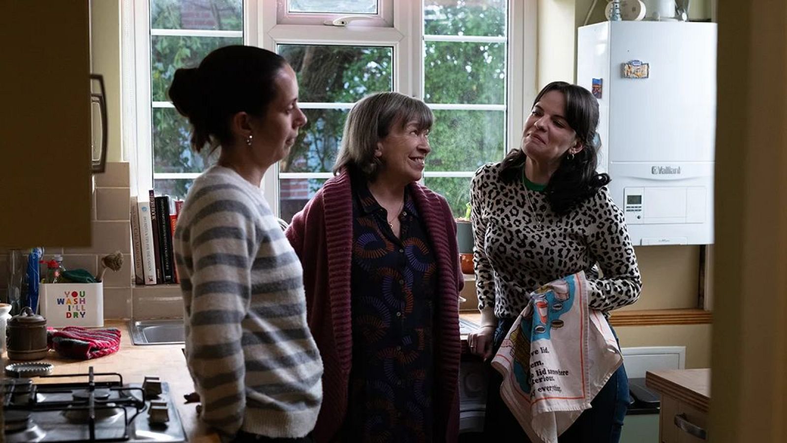 Tres mujeres conversan animadamente en una cocina luminosa. Una sostiene un paño con dibujos, otra sonríe y la tercera mira hacia ellas. Se aprecian detalles como una placa de gas y una caldera Vaillant.