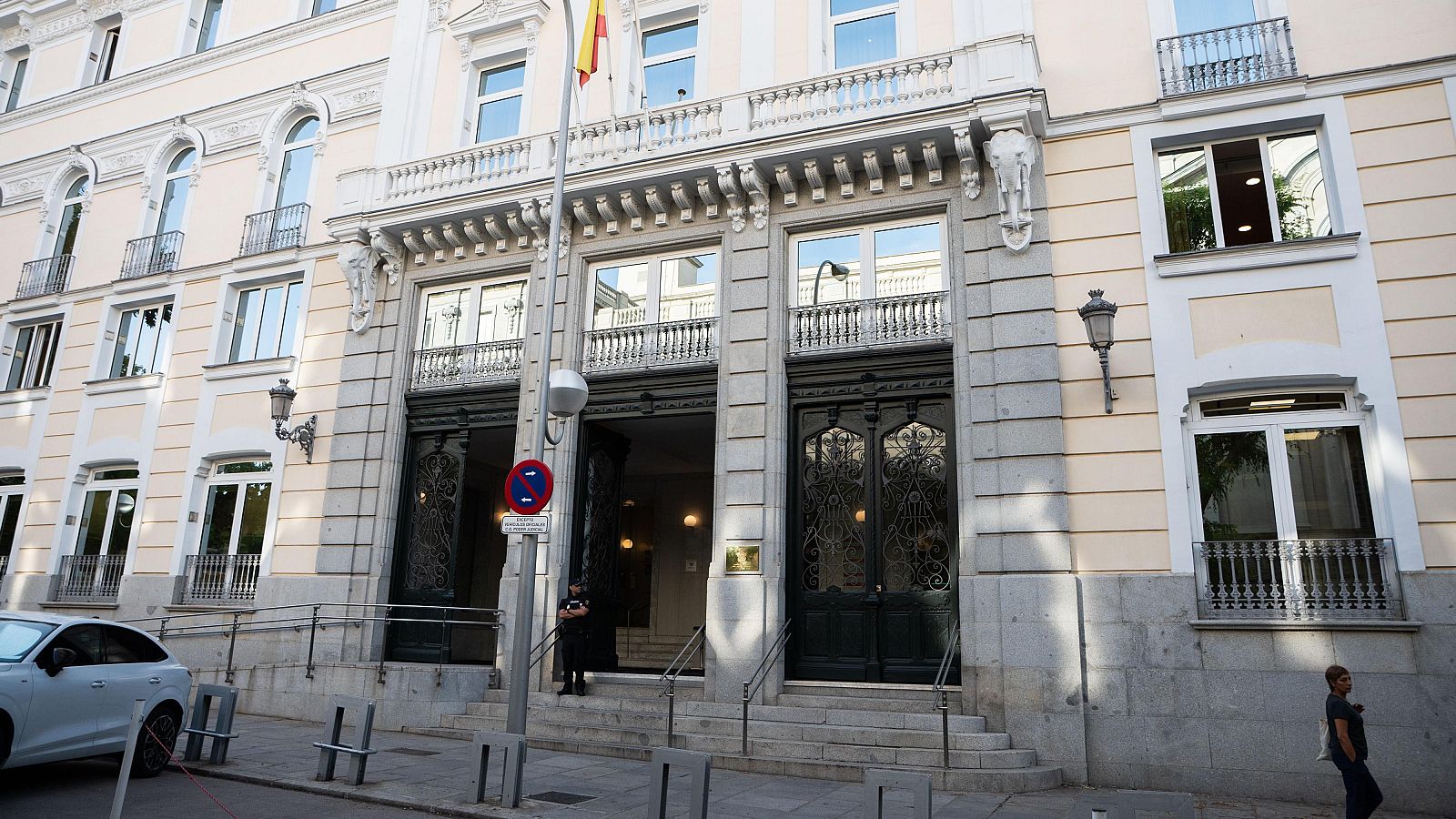 Fachada del CGPJ en Madrid con escalinata, puertas de madera oscura y bandera de España. Señal de 'Prohibido estacionar' y un guardia de seguridad visible.
