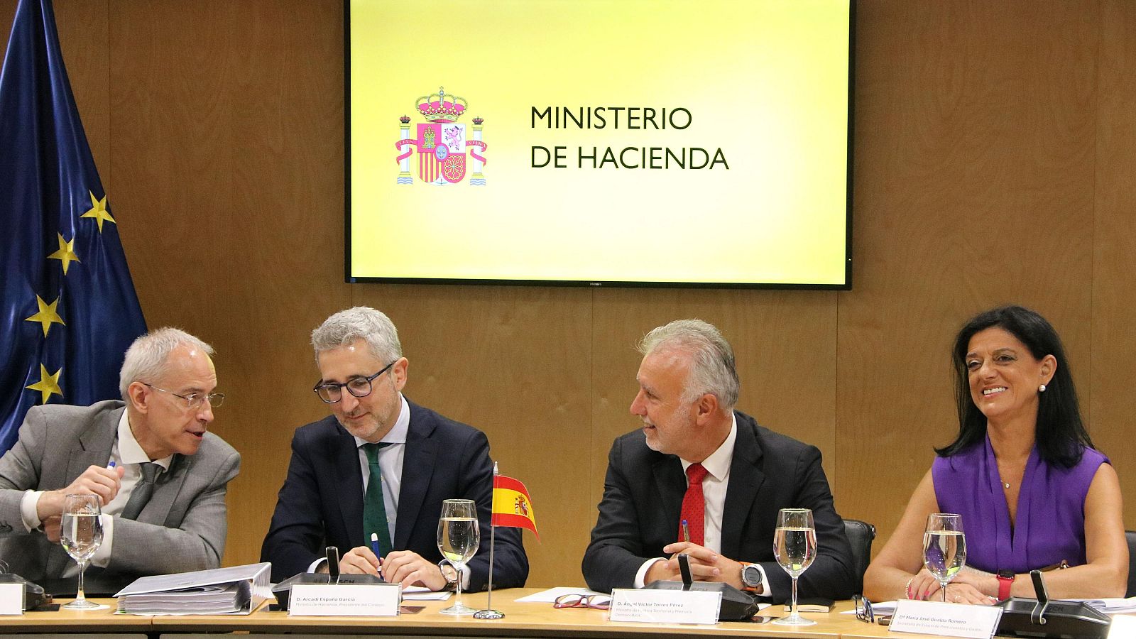 Reunión oficial en el Ministerio de Hacienda con Arcadi España, donde se observan banderas de España y la UE, micrófonos y documentos sobre la mesa.