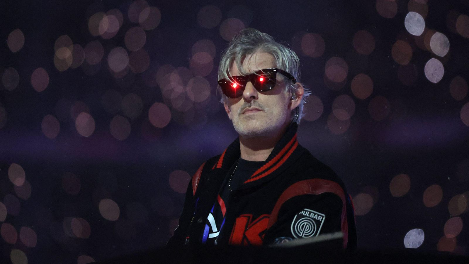 DJ Kavinsky con gafas de sol rojas y chaqueta bomber negra con 'K' blanca y 'PULBAR' en un evento nocturno con luces bokeh.