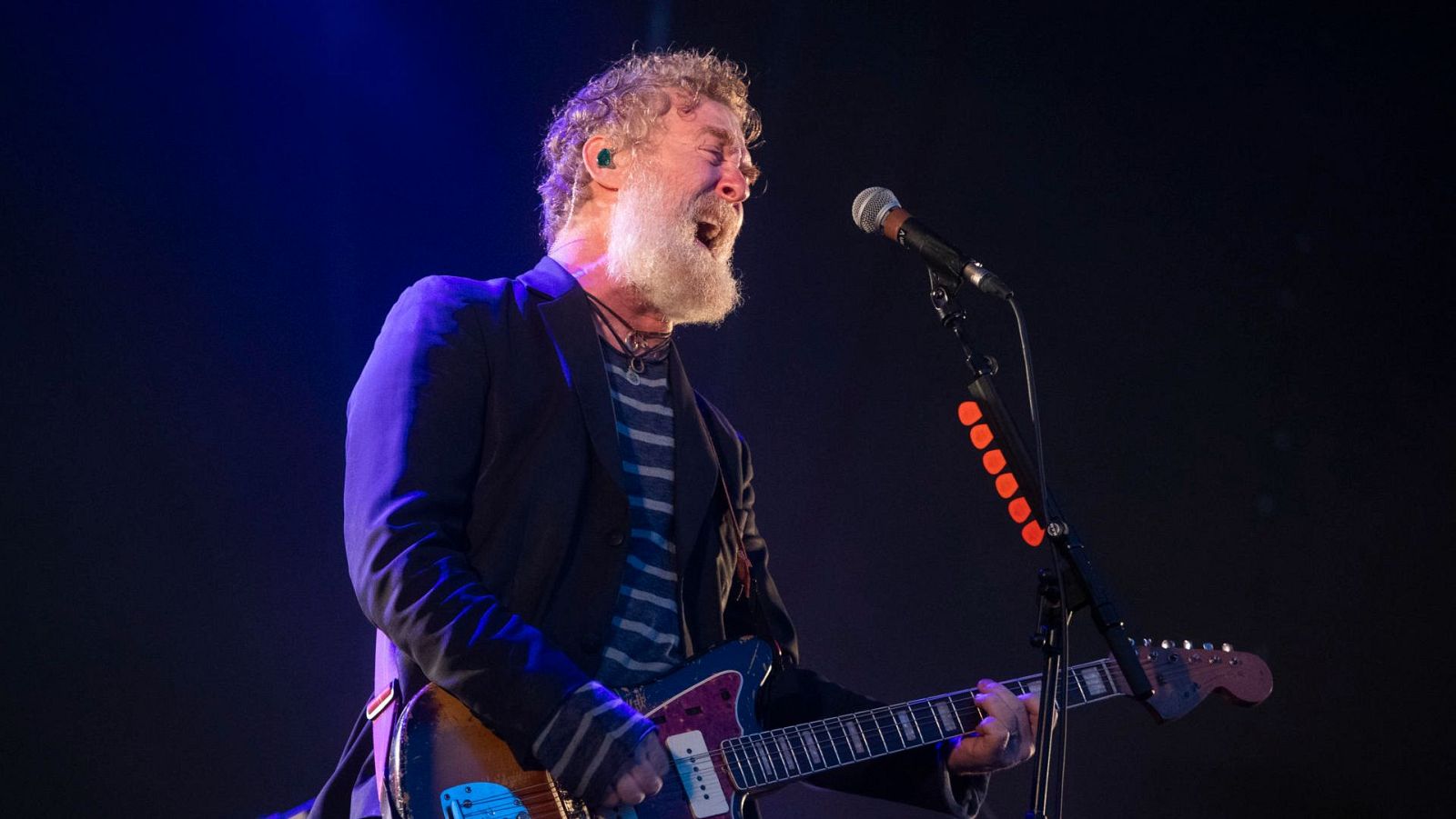Glen Hansard, músico irlandés, canta apasionadamente con los ojos cerrados y la boca abierta, tocando su guitarra eléctrica azul/morada bajo luces de escenario azules y moradas.