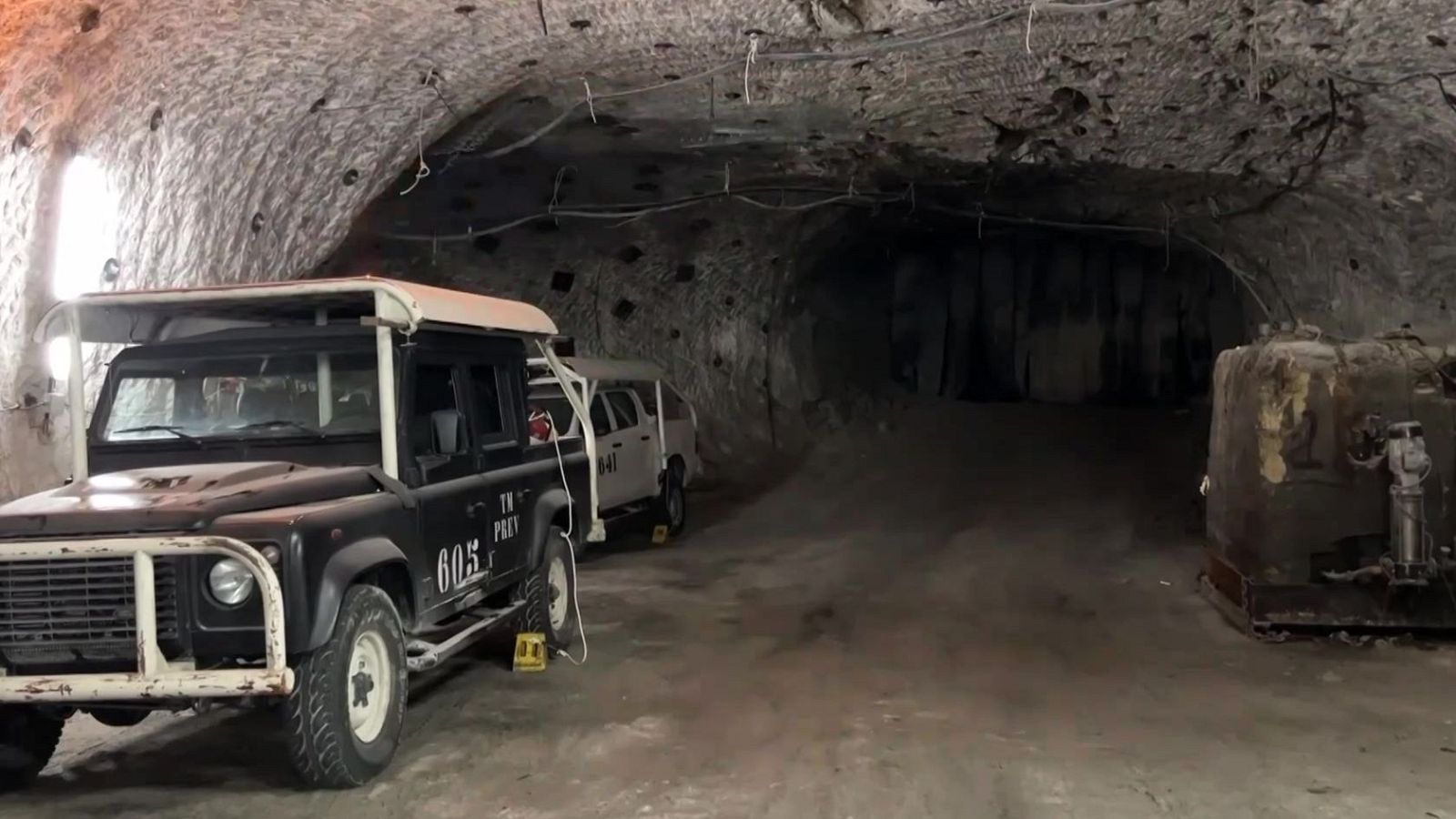Interior de mina con vehículo utilitario "TK PREV 605" y maquinaria industrial. Túnel rocoso con iluminación tenue.