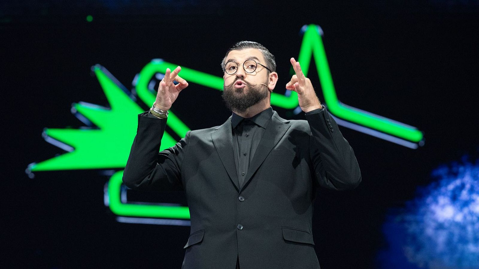 Manu Sánchez, con traje oscuro y gafas, gesticula en un escenario iluminado por formas verdes neón, durante una presentación.