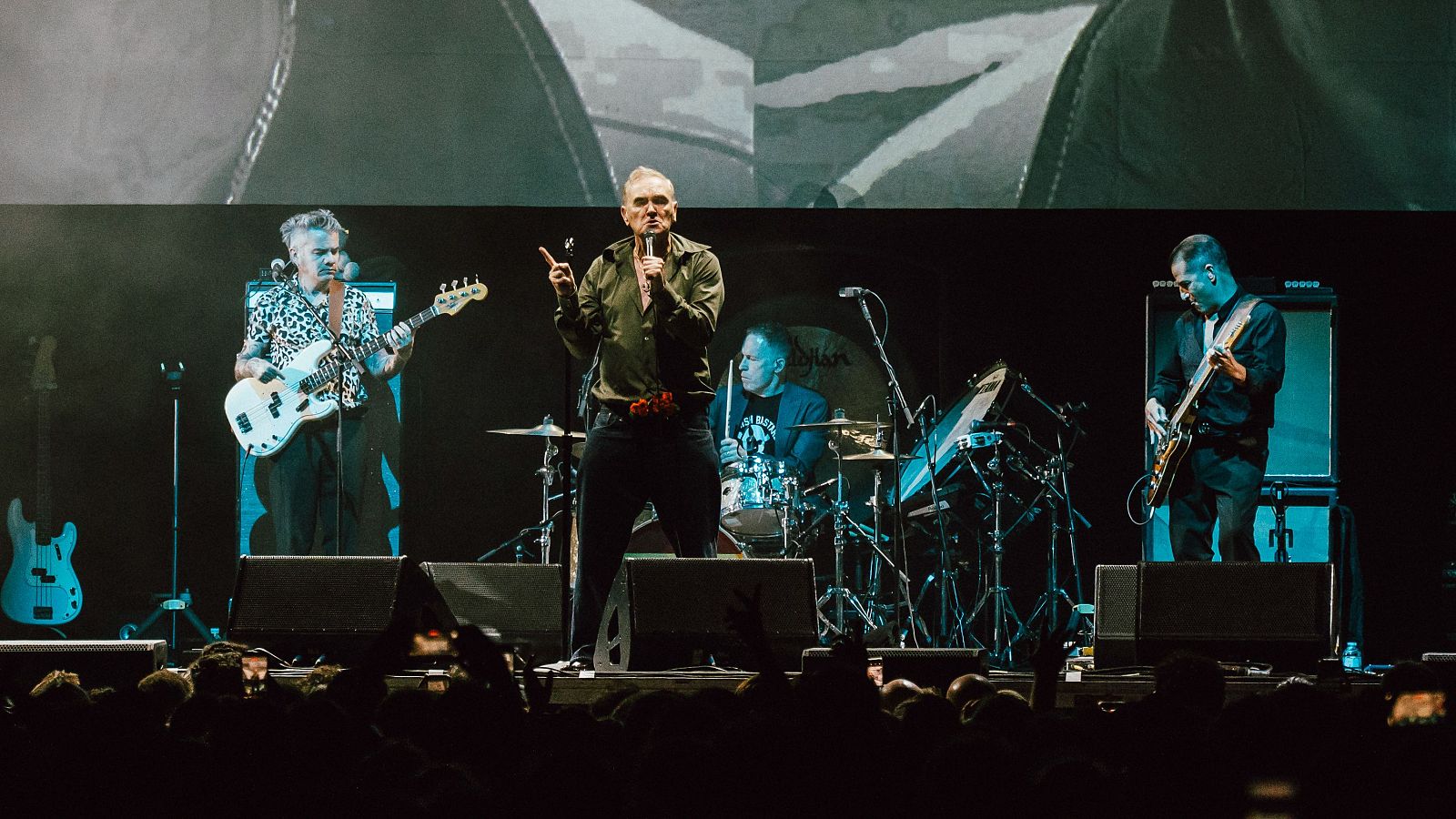 Morrissey en concierto en Madrid, cantando al micrófono con su banda. Escenario con pantalla abstracta y público al frente.