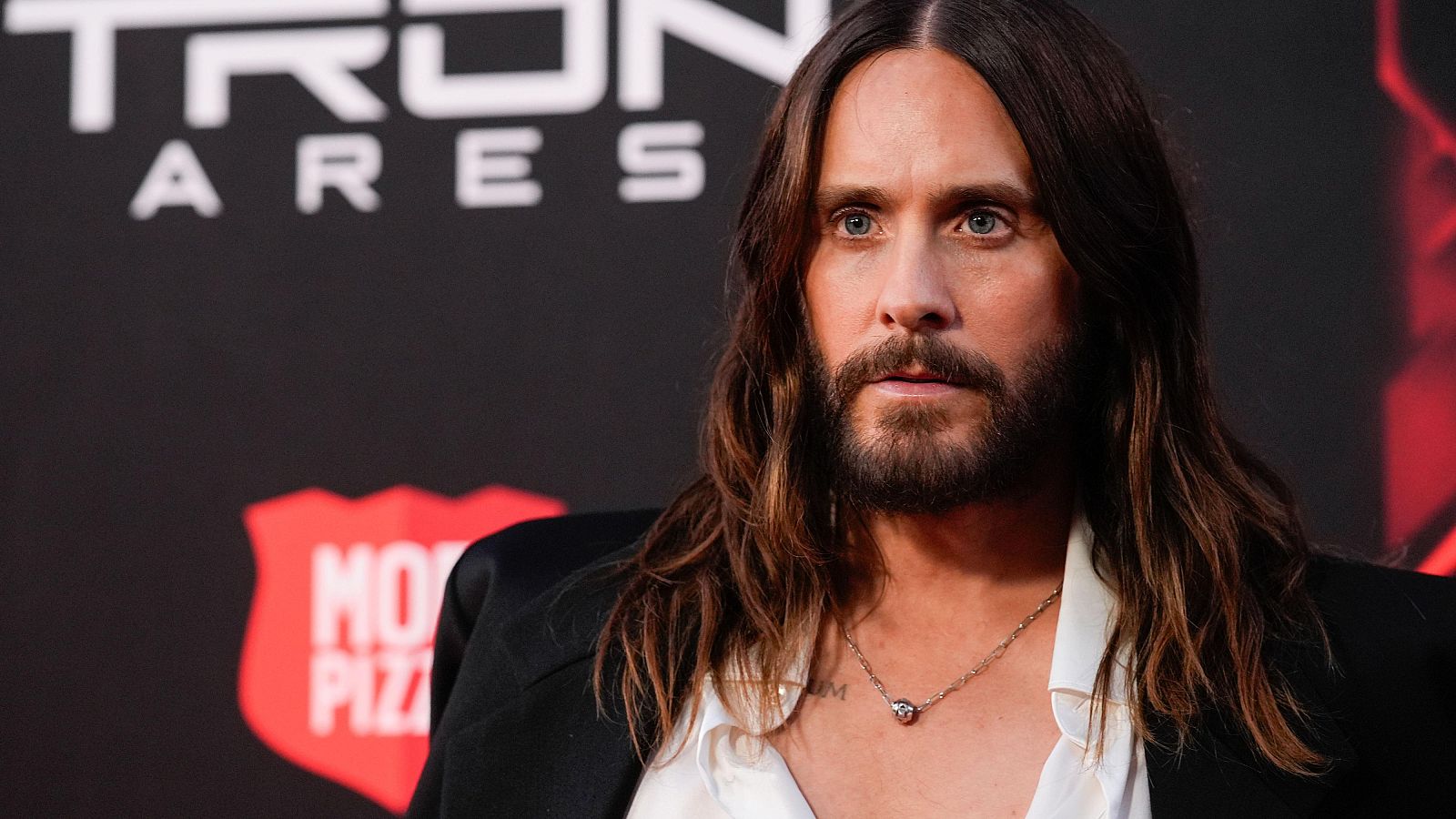 Primer plano de Jared Leto con cabello largo y barba, vestido con chaqueta oscura y collar. Fondo con "TRON ARES" y logo parcial.