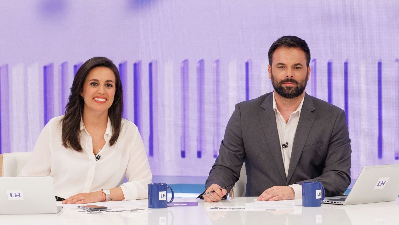 Aida Bao y Álex Barreiro, presentadores de 'La hora de La 1', posan en un set de televisión con ordenadores, tazas y papeles sobre una mesa blanca.