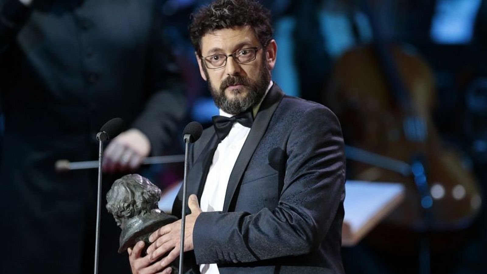Manolo Solo, actor español, con su Premio Goya en un evento de gala. Lleva esmoquin y se aprecia un micrófono frente a él.