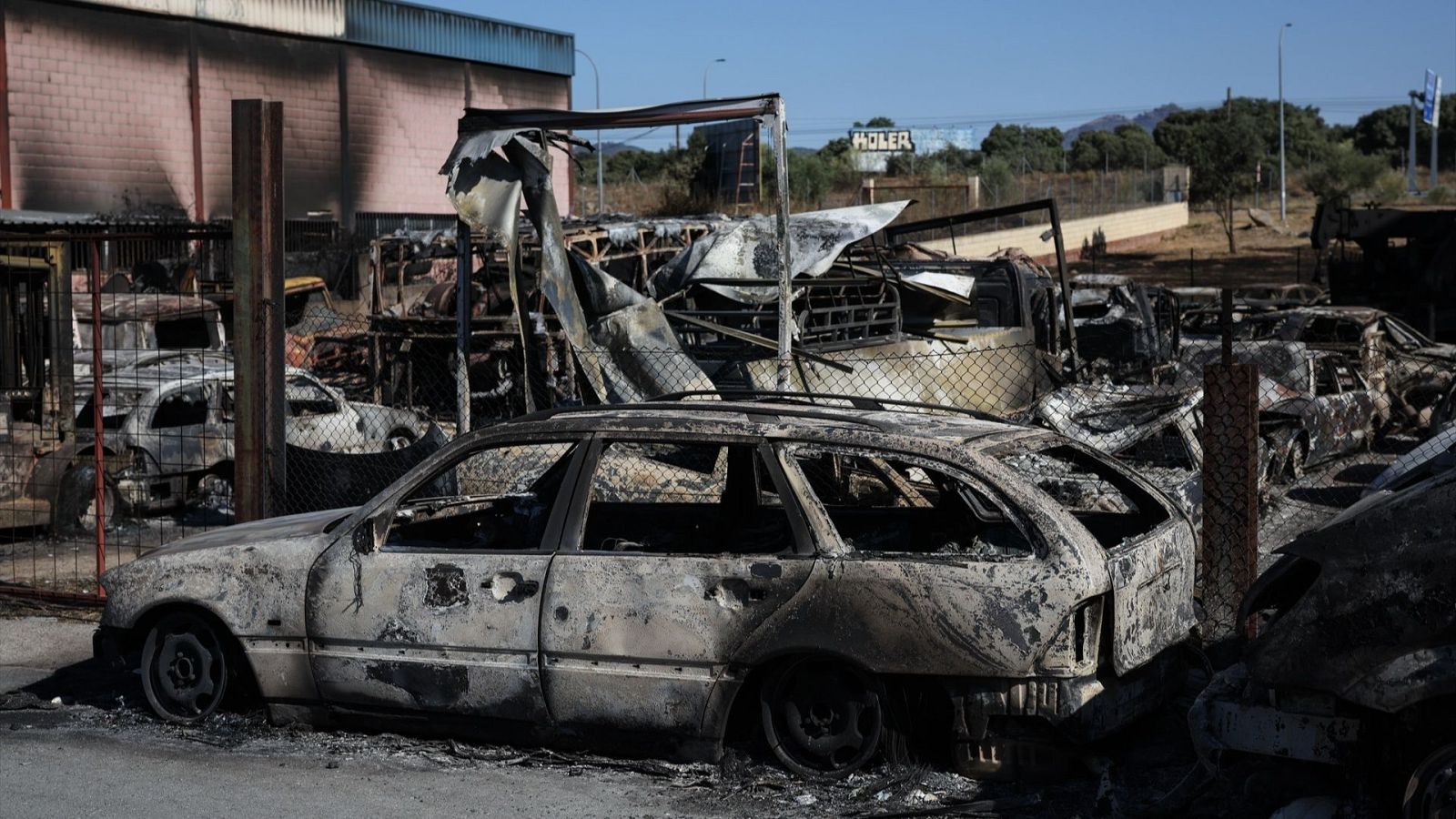 Vehículos calcinados y estructuras dañadas por un incendio, con un cartel "HOLER" al fondo, bajo un cielo azul.