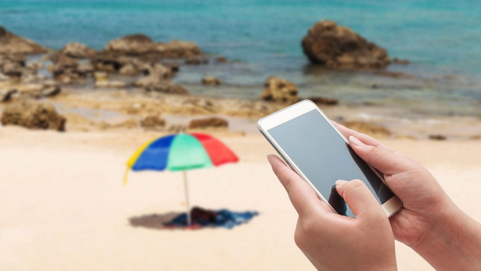 Manos sujetan un smartphone en una playa soleada, ilustrando la dificultad de desconectar en vacaciones.