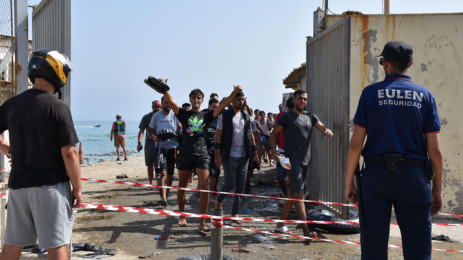 Hombres cruzan la frontera en Ceuta, algunos descalzos y empapados. Un guardia de EULEN SEGURIDAD y un hombre con logo Adidas son visibles.