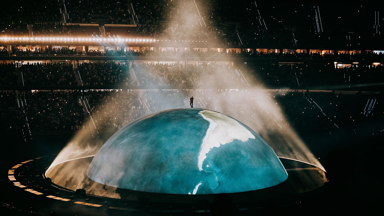 Kanye West actuando en el Estadio Metropolitano de Madrid sobre un escenario de globo terráqueo, rodeado de humo y luces, ante una multitud.