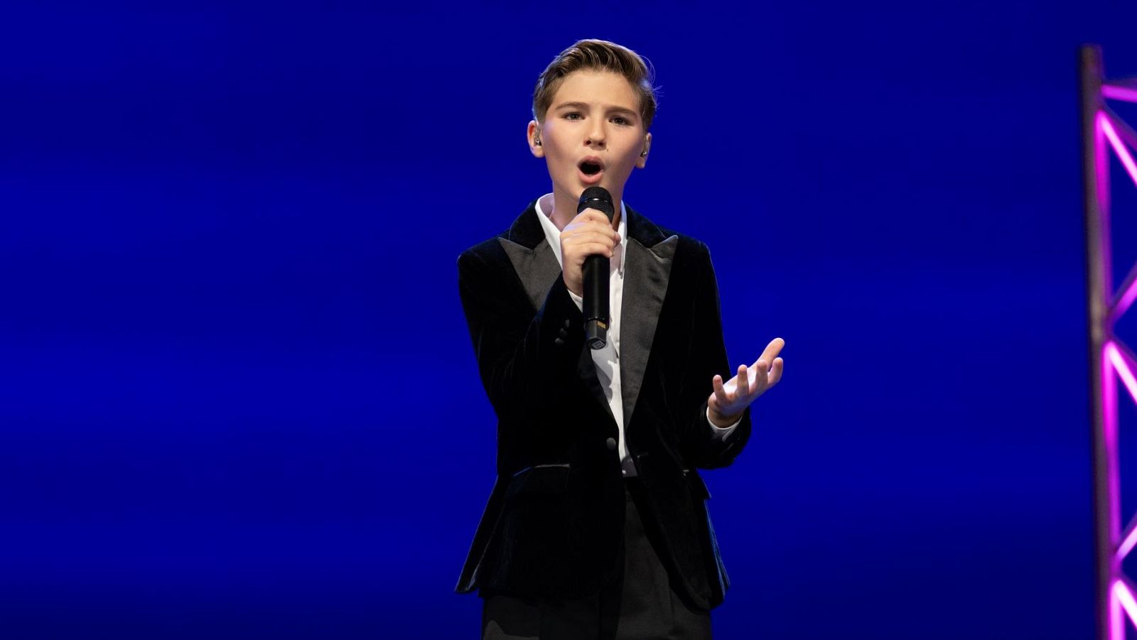Lucas Paulano, representante de España en Eurovisión Junior, canta con un micrófono, vestido de traje negro de terciopelo y camisa blanca, con la mano izquierda levantada en un gesto expresivo, sobre un fondo azul con luces rosas.