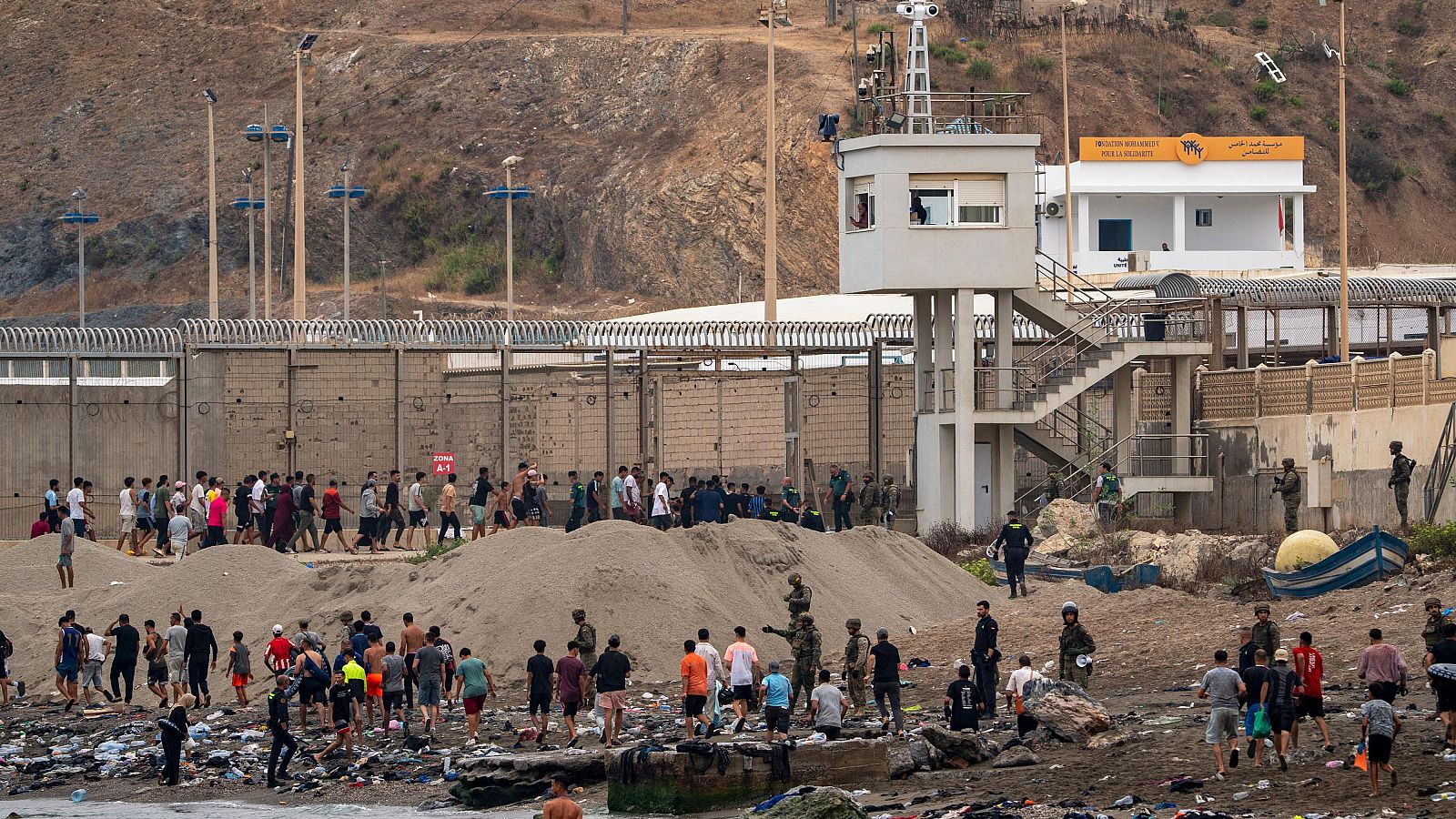 Un puesto de seguridad fronteriza marroquí con el letrero "MOROCCAN BORDER SECURITY" y "CUSTOMS" supervisa a grupos de hombres jóvenes en una playa con basura.
