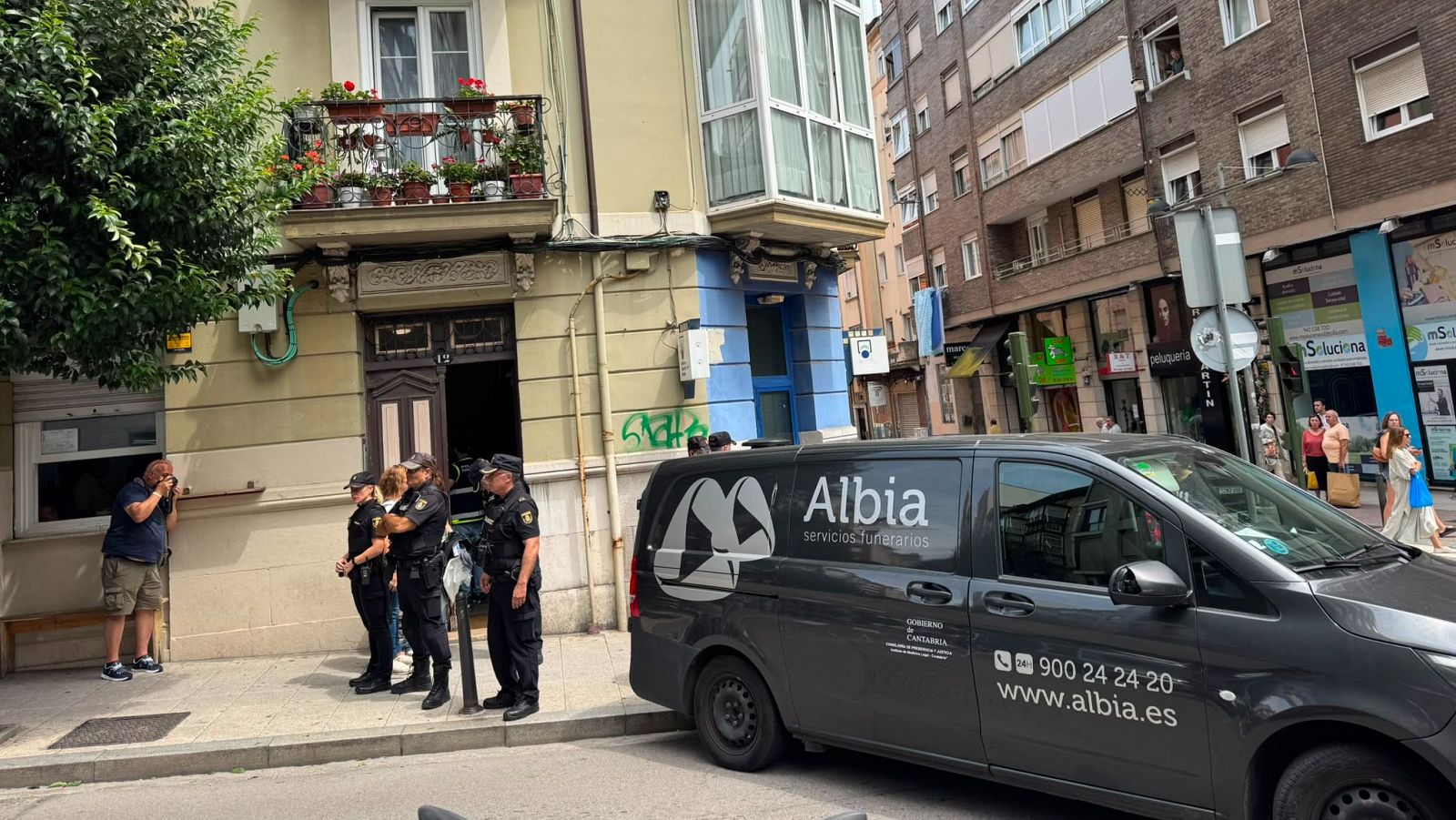 Furgón funerario "Albia servicios funerarios" de Mercedes-Benz en calle de Cantabria, rodeado de policías y un civil documentando la escena.
