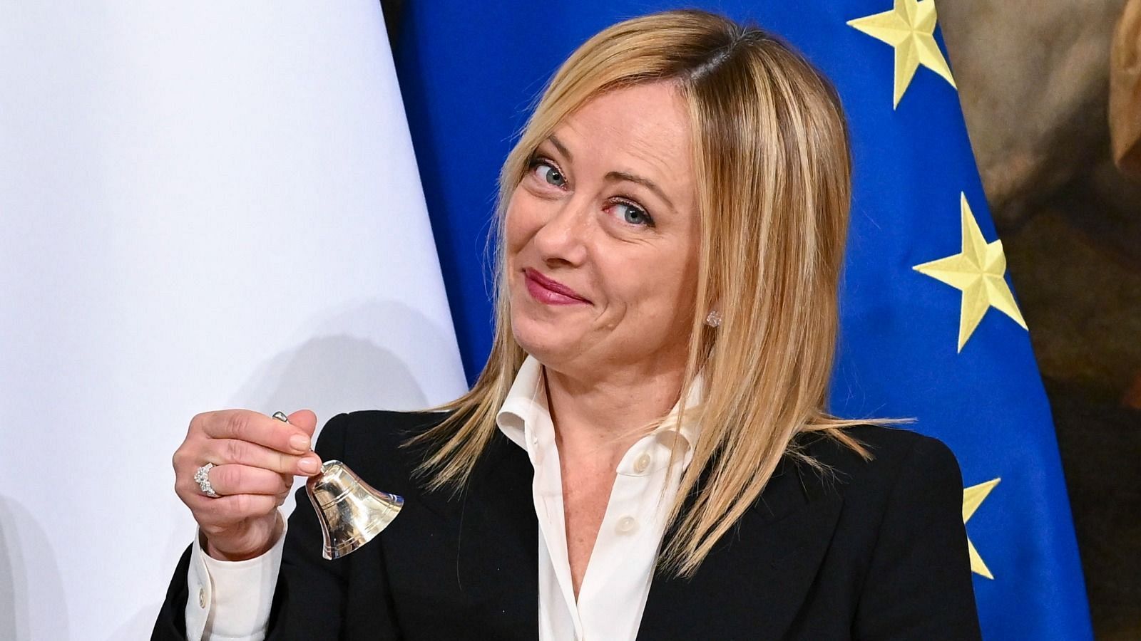 Giorgia Meloni, con chaqueta negra y camisa blanca, sonríe mientras sostiene una campana dorada. Detrás de ella, se observa la bandera de la Unión Europea.