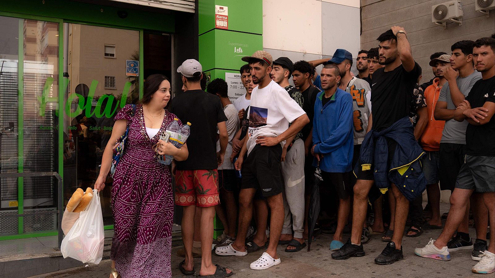 Mujer con vestido morado y blanco camina con pan y agua. Multitud de hombres jóvenes espera frente a edificio con "Hola" y horarios 7:00-22:00.
