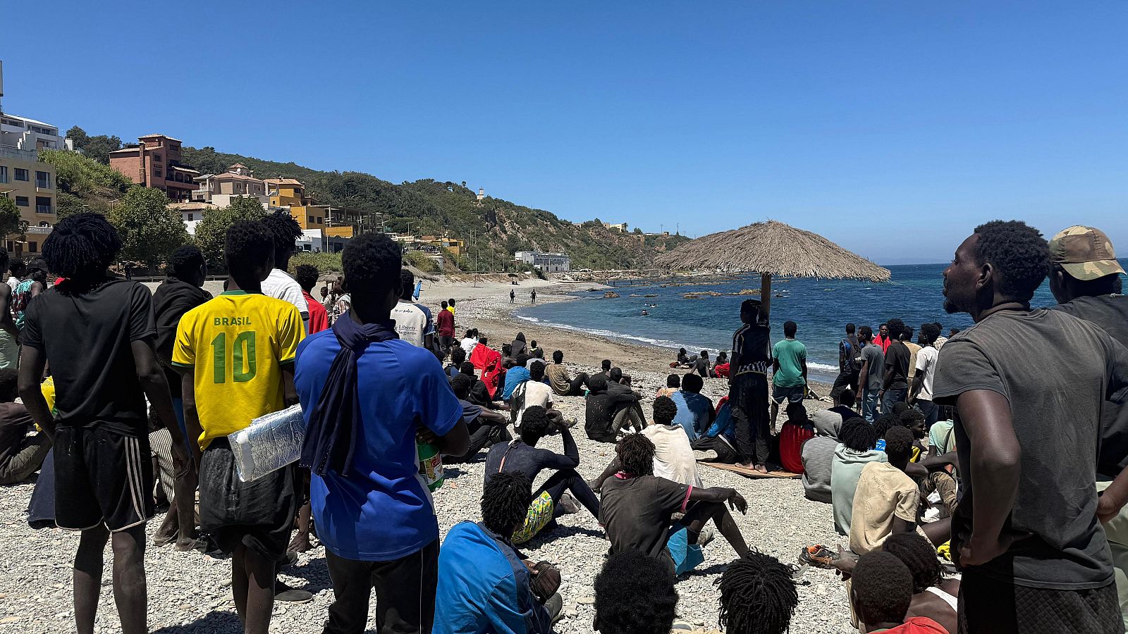 Playa soleada con multitud de hombres de piel oscura, uno con camiseta de Brasil '10'. Mar azul y colinas verdes al fondo.