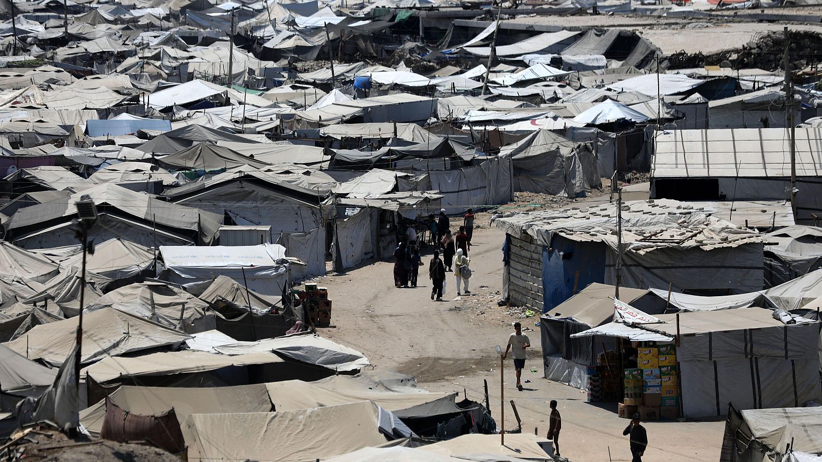 Vista panorámica de un extenso campamento de tiendas de campaña y refugios improvisados en terreno árido, con personas transitando y estructuras de ayuda humanitaria y venta.