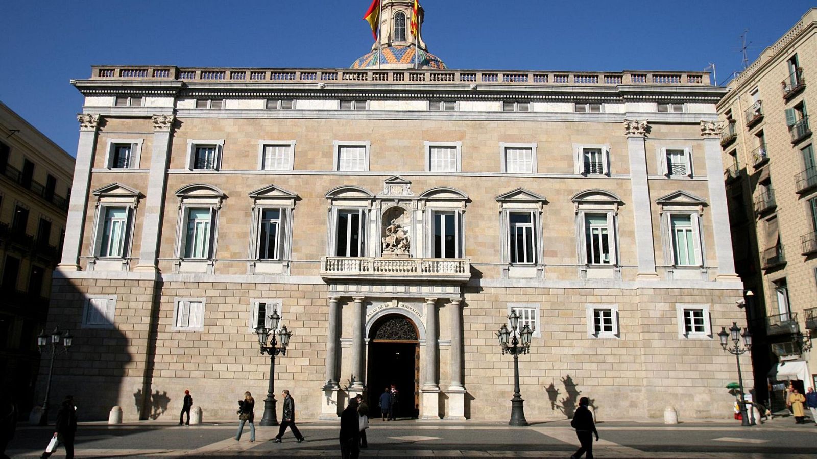 Façana del Palau de la Generalitat de Catalunya amb escultura de Sant Jordi, cúpula amb mosaic i bandera d'Espanya. Plaça amb faroles i vianants.