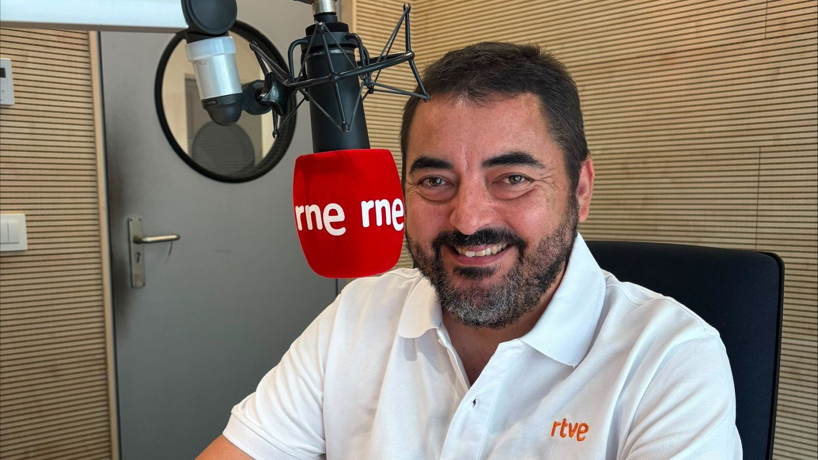 Periodista español Pablo López sonríe a cámara en estudio de radio, vistiendo camiseta blanca con logo RTVE y micrófono "rne rne" a su lado. Fondo de lamas de madera.