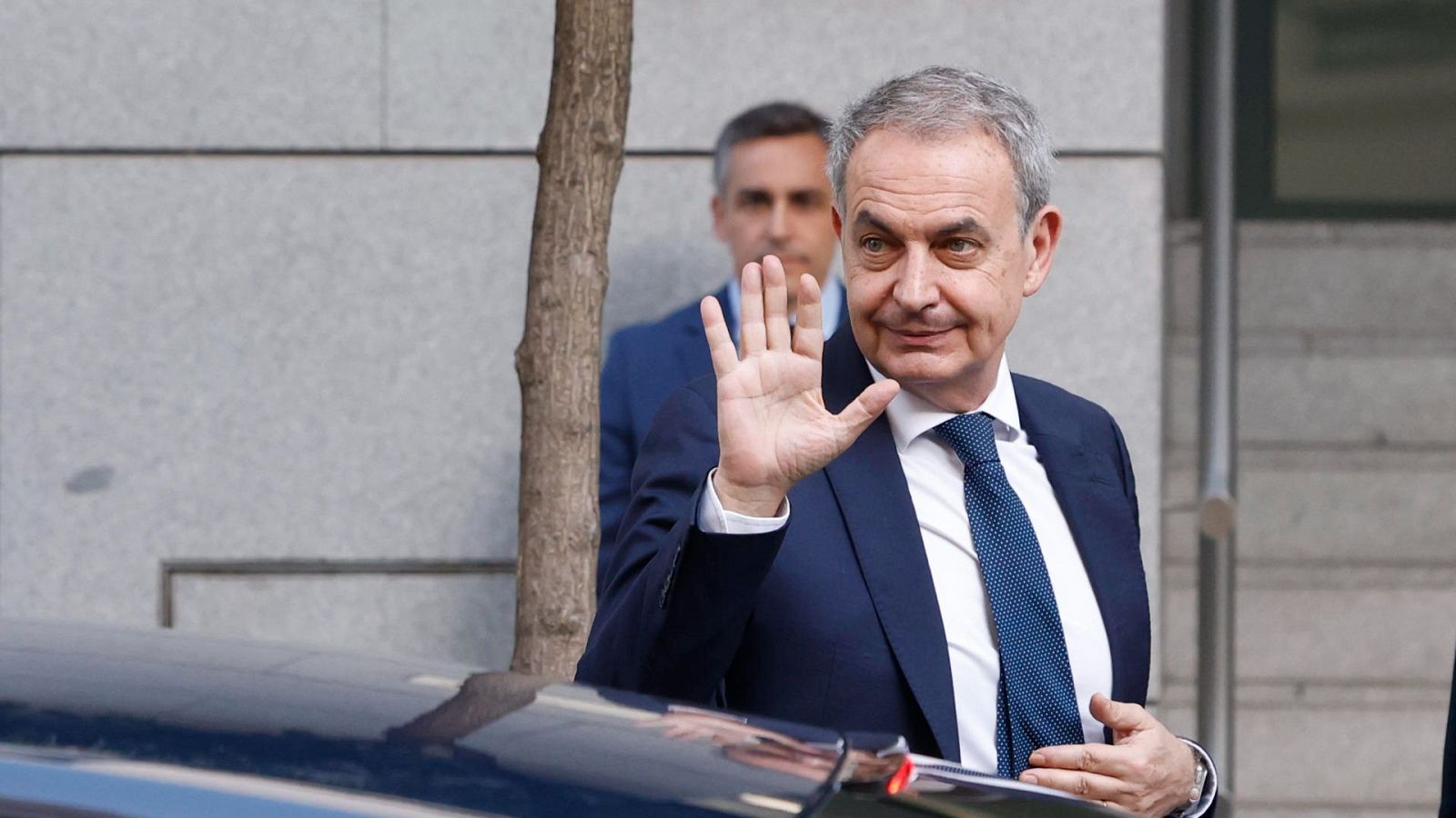 José Luis Rodríguez Zapatero saluda con la mano derecha levantada, vistiendo traje azul marino. Detrás, un hombre con traje oscuro y un árbol. Fondo de edificio moderno y coche.