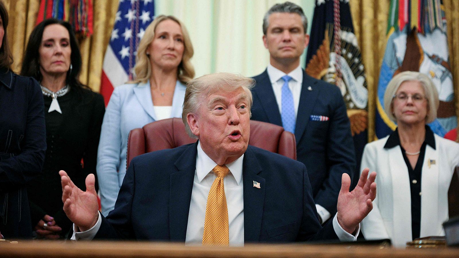 Donald Trump en el Despacho Oval, con la boca abierta y manos levantadas, rodeado de cinco personas y dos banderas.