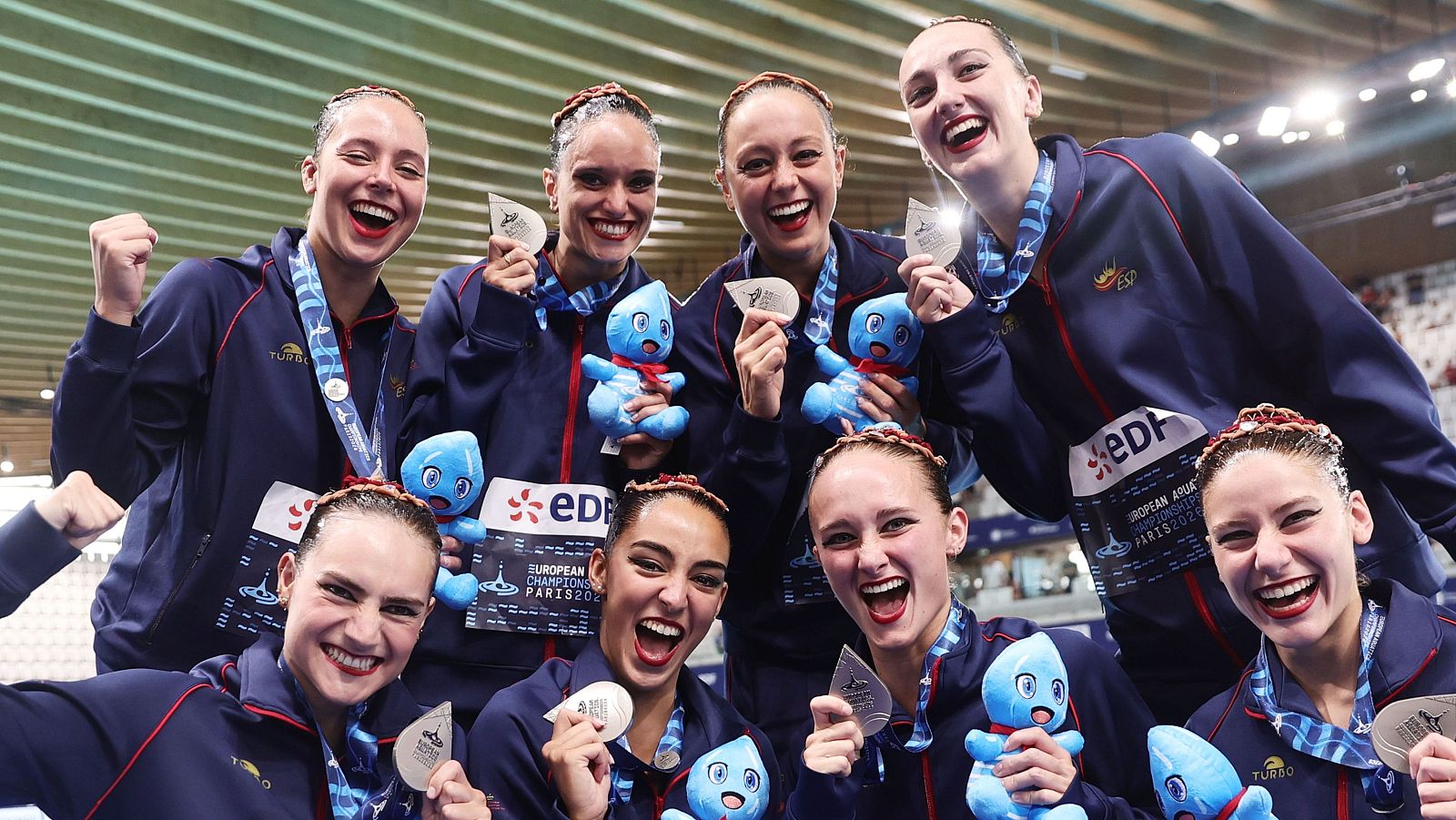 Nadadoras artísticas españolas celebran medallas de plata en París, luciendo chaquetas con 'ESP' y sosteniendo mascotas del evento. Se distingue el logo 'EDP' y 'EUROPEAN ARTISTIC SWIMMING CHAMPIONSHIPS PARIS 202...'.